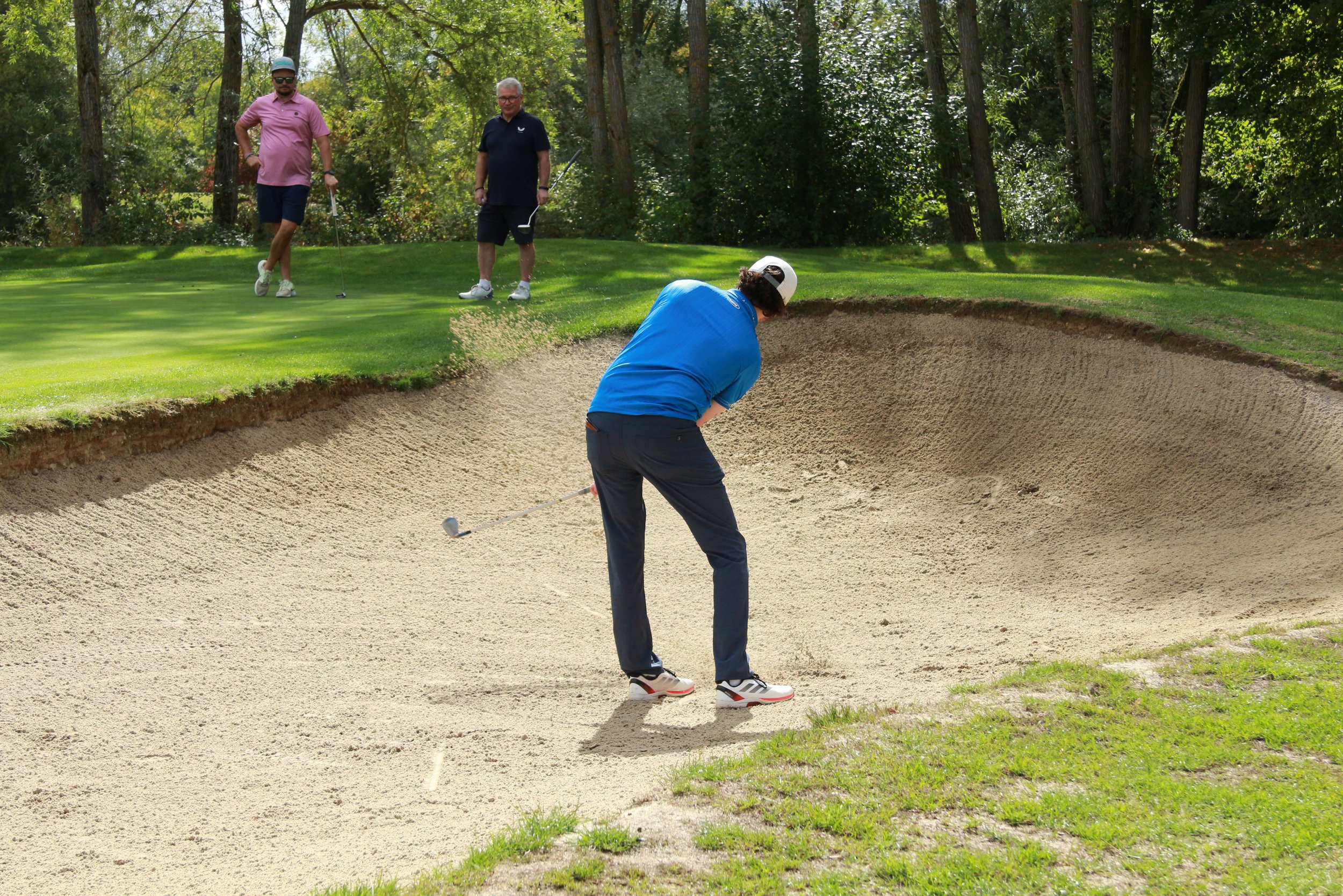 Three men playing golf on a golf course, with two of them near the green and the third in a sand bunker preparing to hit the ball.