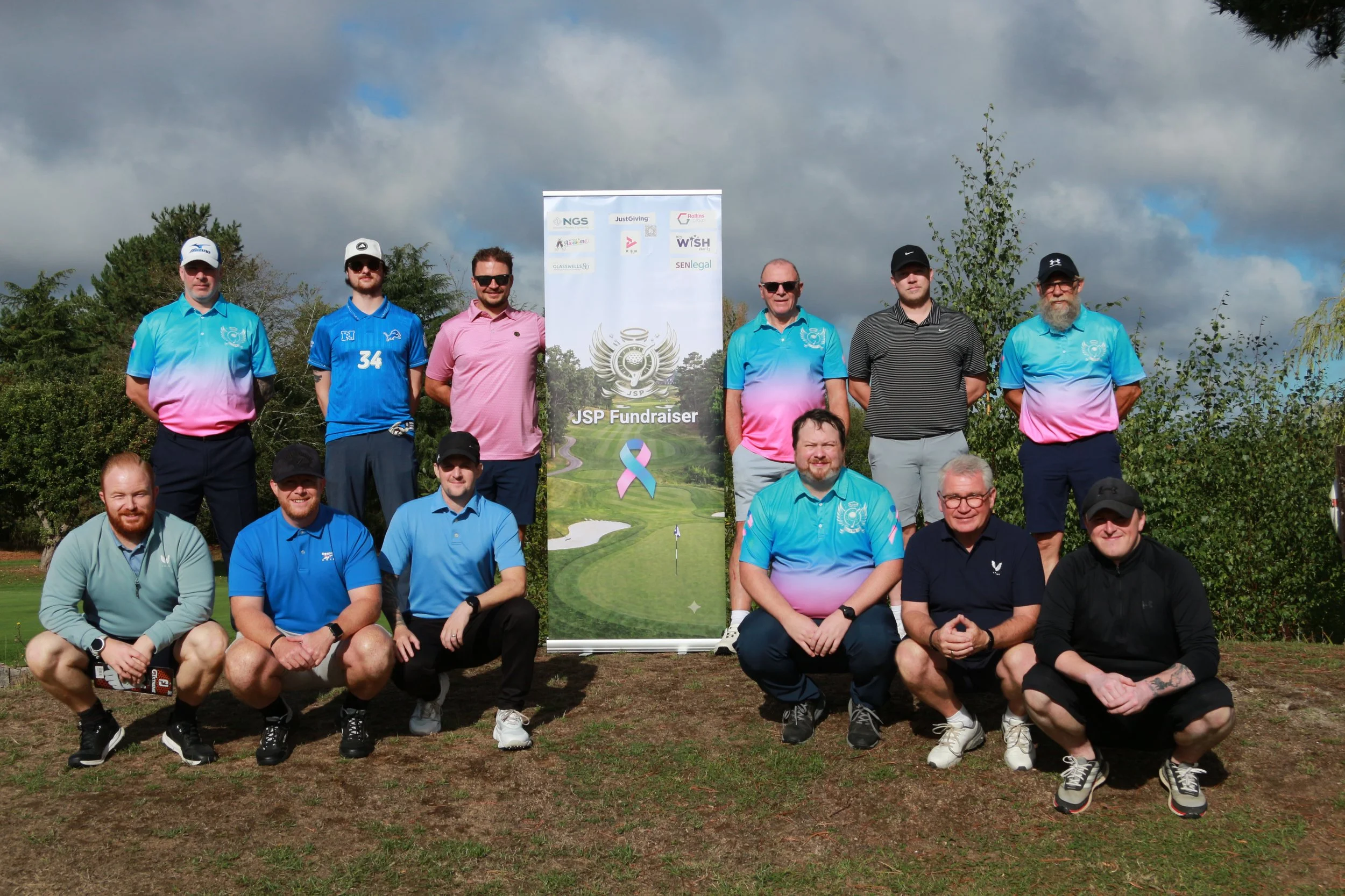 Group of men posing outdoors in front of a banner for a fundraising event called JSP Fundraiser, with trees and cloudy sky in the background.