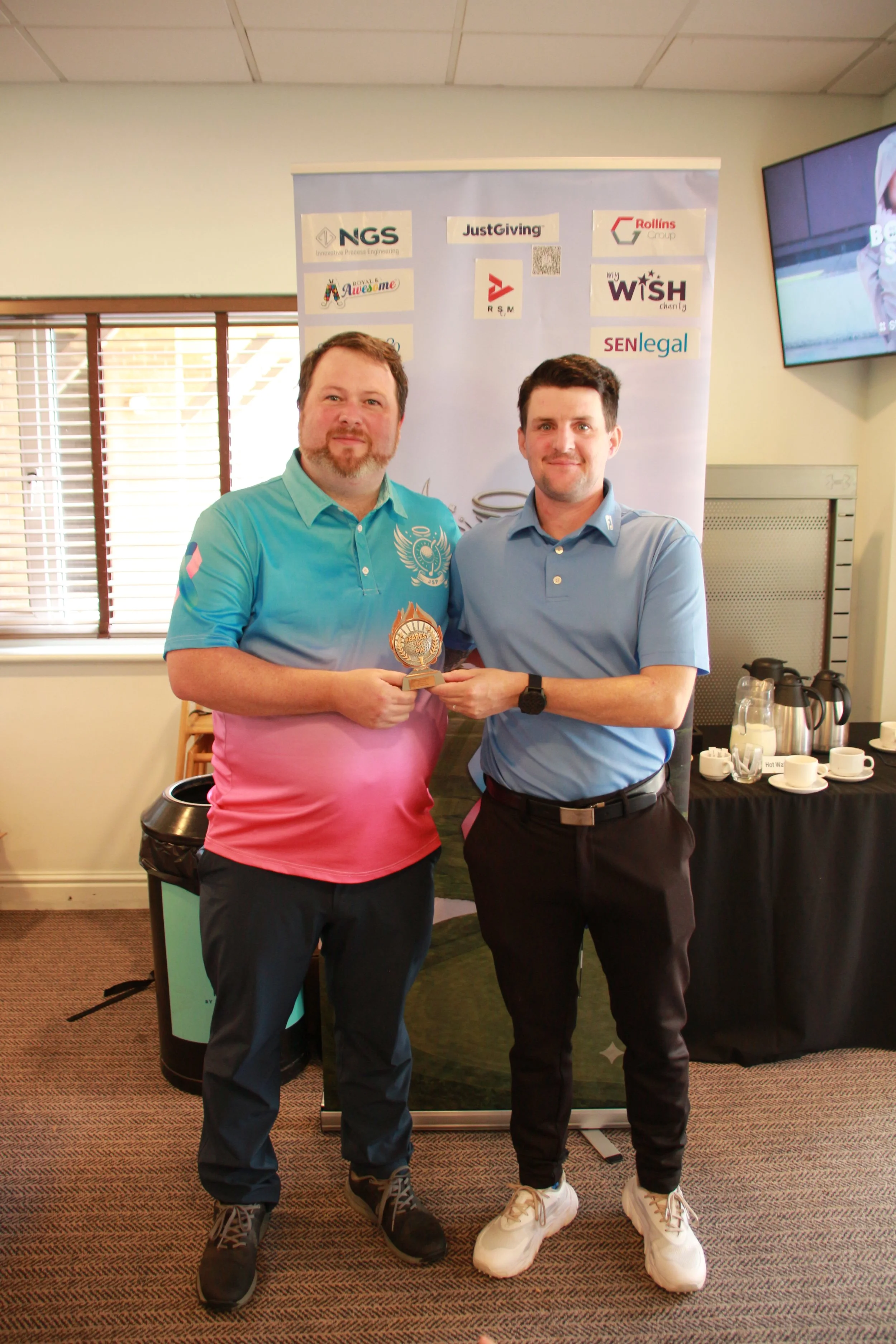 Two men standing indoors, holding a trophy, in front of a banner with various company logos, including 'NGS,' 'JustGiving,' 'Rollins Group,' 'myWISH,' and 'SENlegal.' One man is wearing a light blue polo shirt, and the other is wearing a darker blue 