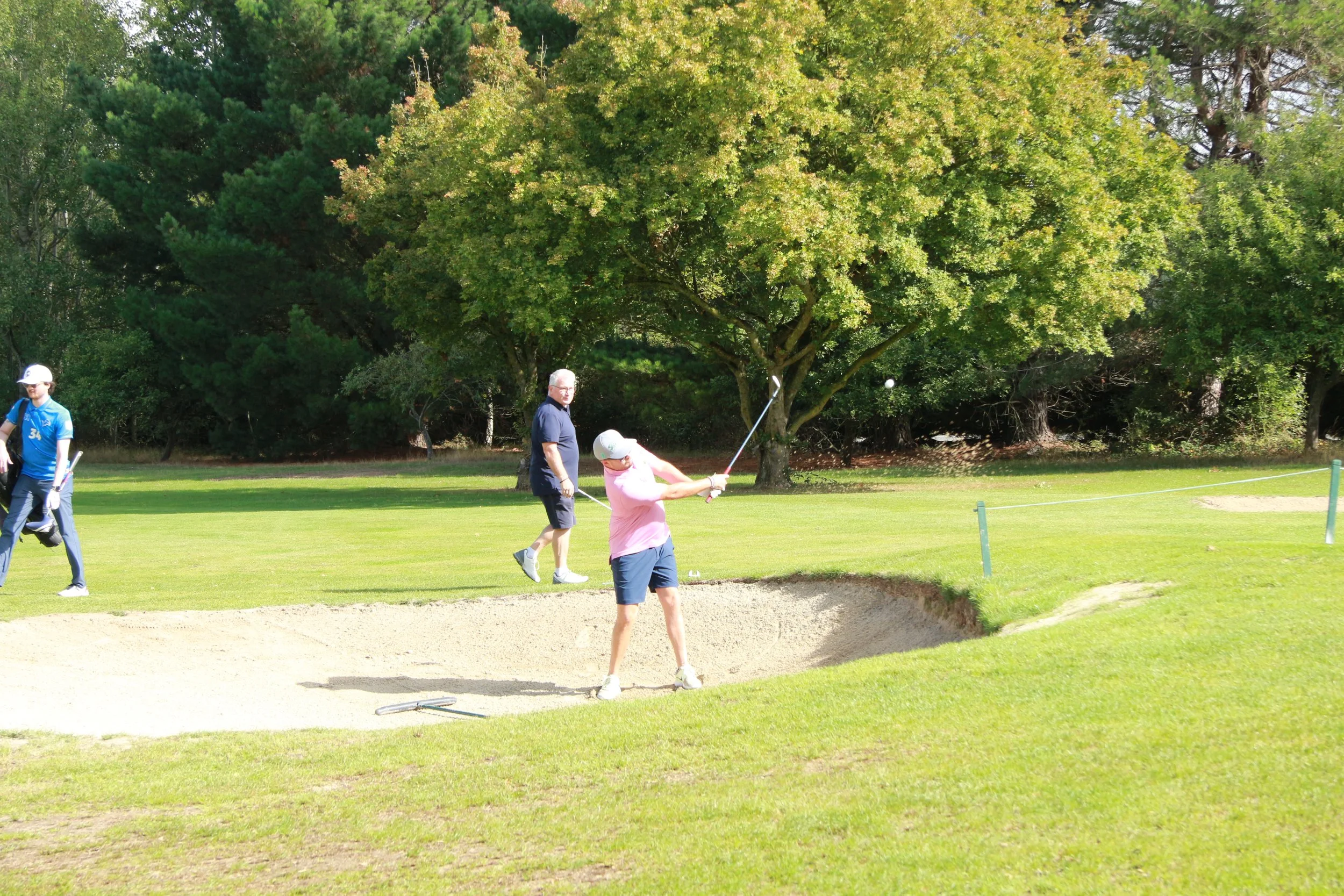 People playing golf on a course, with one person in pink making a shot from a sand trap, and others watching nearby, surrounded by green trees.