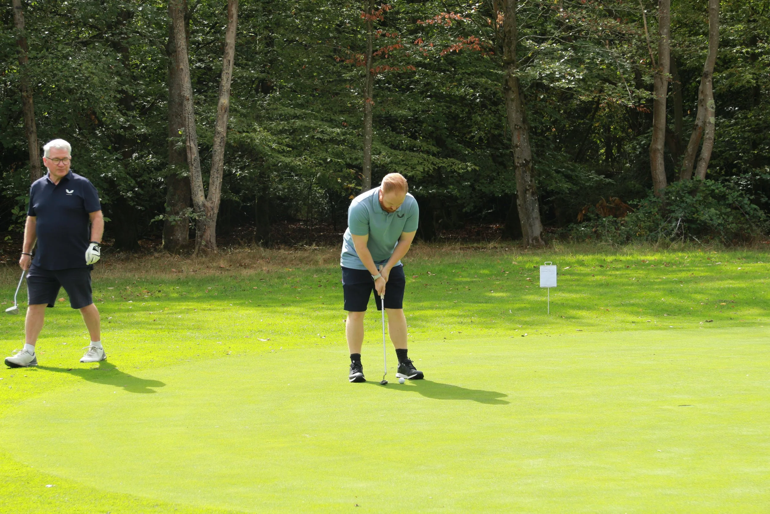 Two men playing golf on a green with a wooded background. One man is standing with a golf club, and the other is preparing to putt the golf ball.