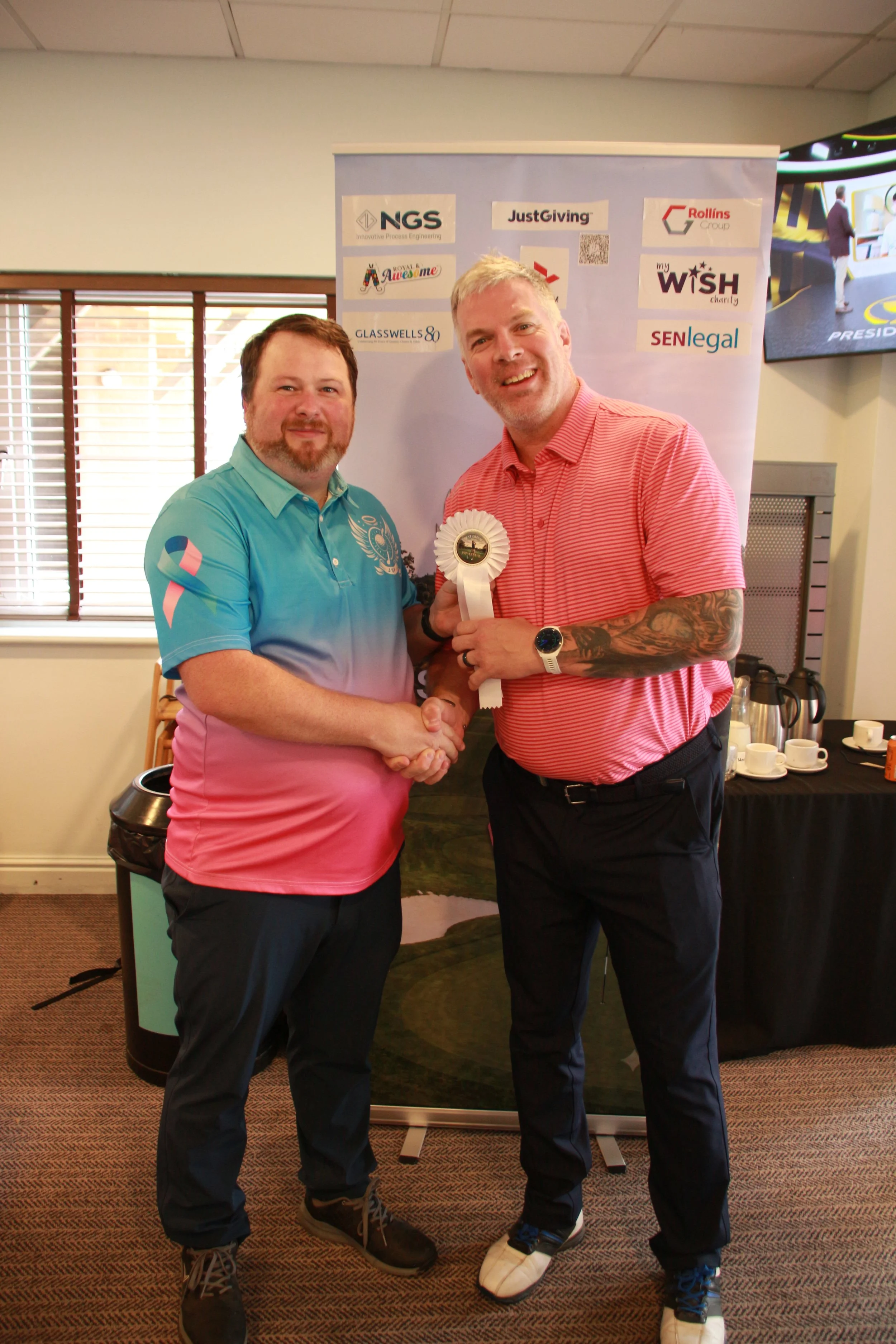 Two men shaking hands and smiling at an awards ceremony. One man is wearing a pink and blue polo shirt, and the other is in a red striped polo shirt, holding a white ribbon. Behind them are logos of various sponsors on a banner, a window with blinds,
