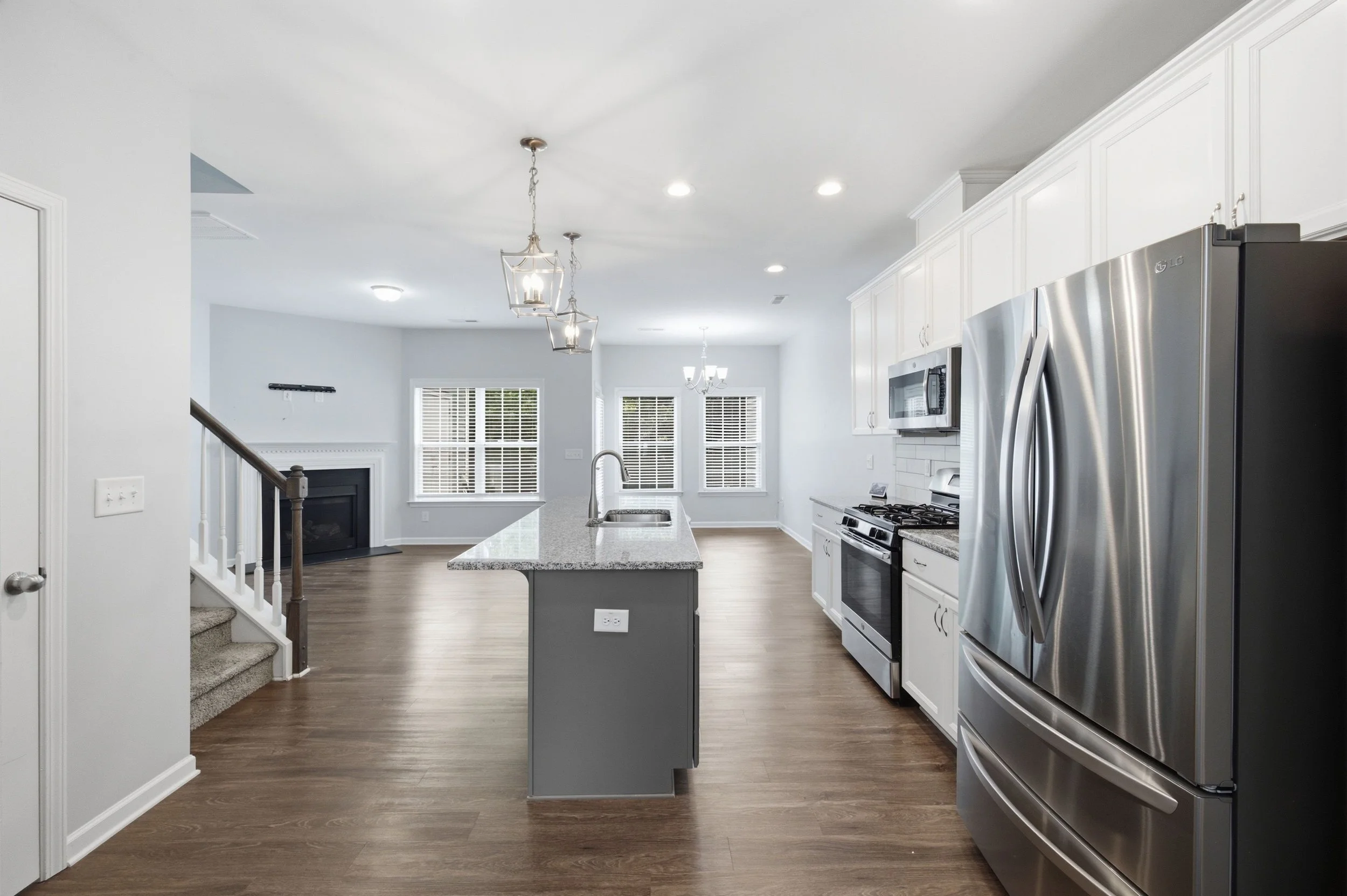 Modern kitchen with stainless steel appliances, white cabinets, granite countertops, and hardwood floors. A kitchen island is in the center, with hanging pendant lights above. Windows with blinds allow natural light to enter. BitBrick Cleveland.