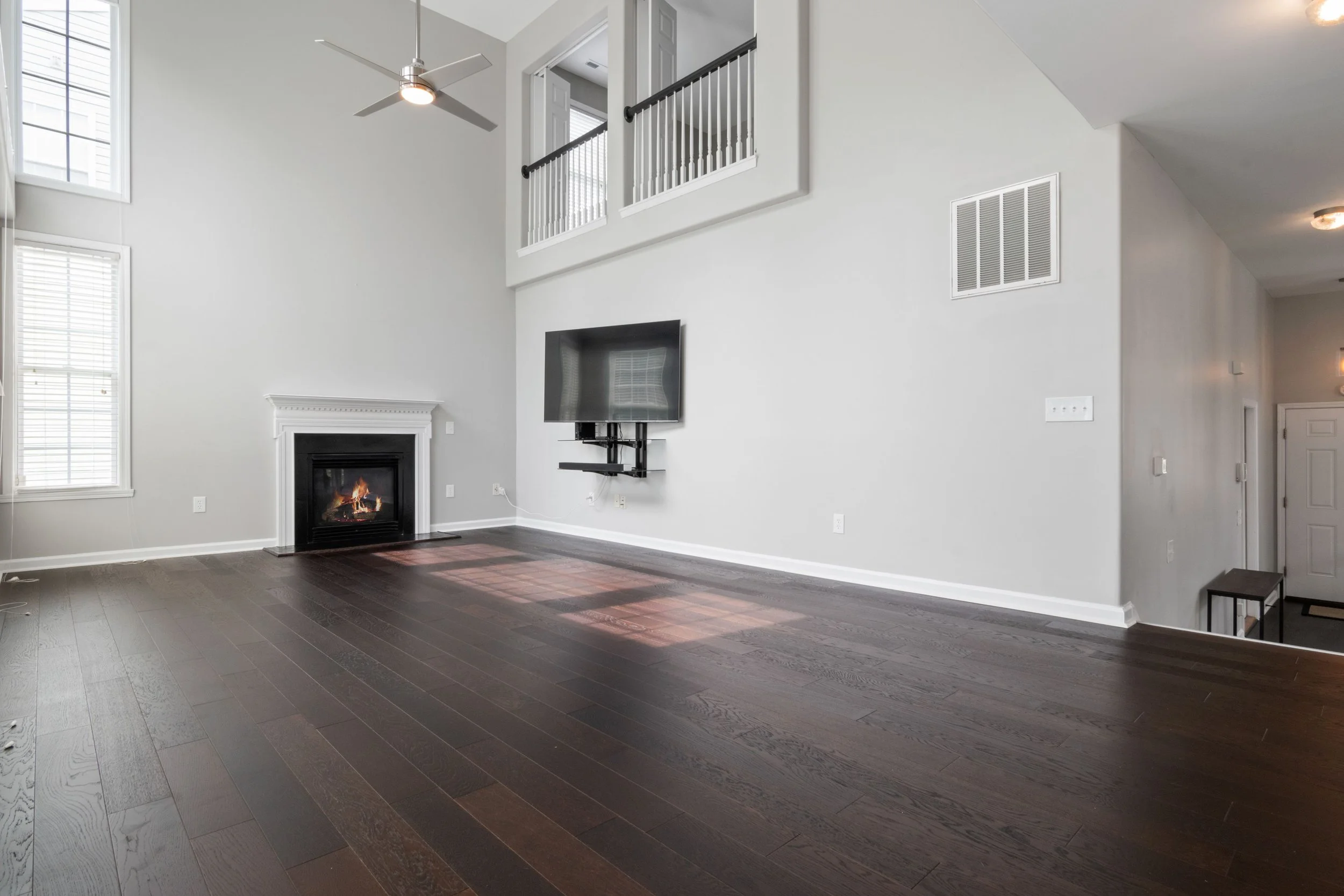 Empty living room with high vaulted ceiling, hardwood floors, fireplace, wall-mounted flat-screen TV, large windows, and a balcony with white railing. BitBrick - Cleveland's local team for flooring, kitchen and bath remodels, and general construction