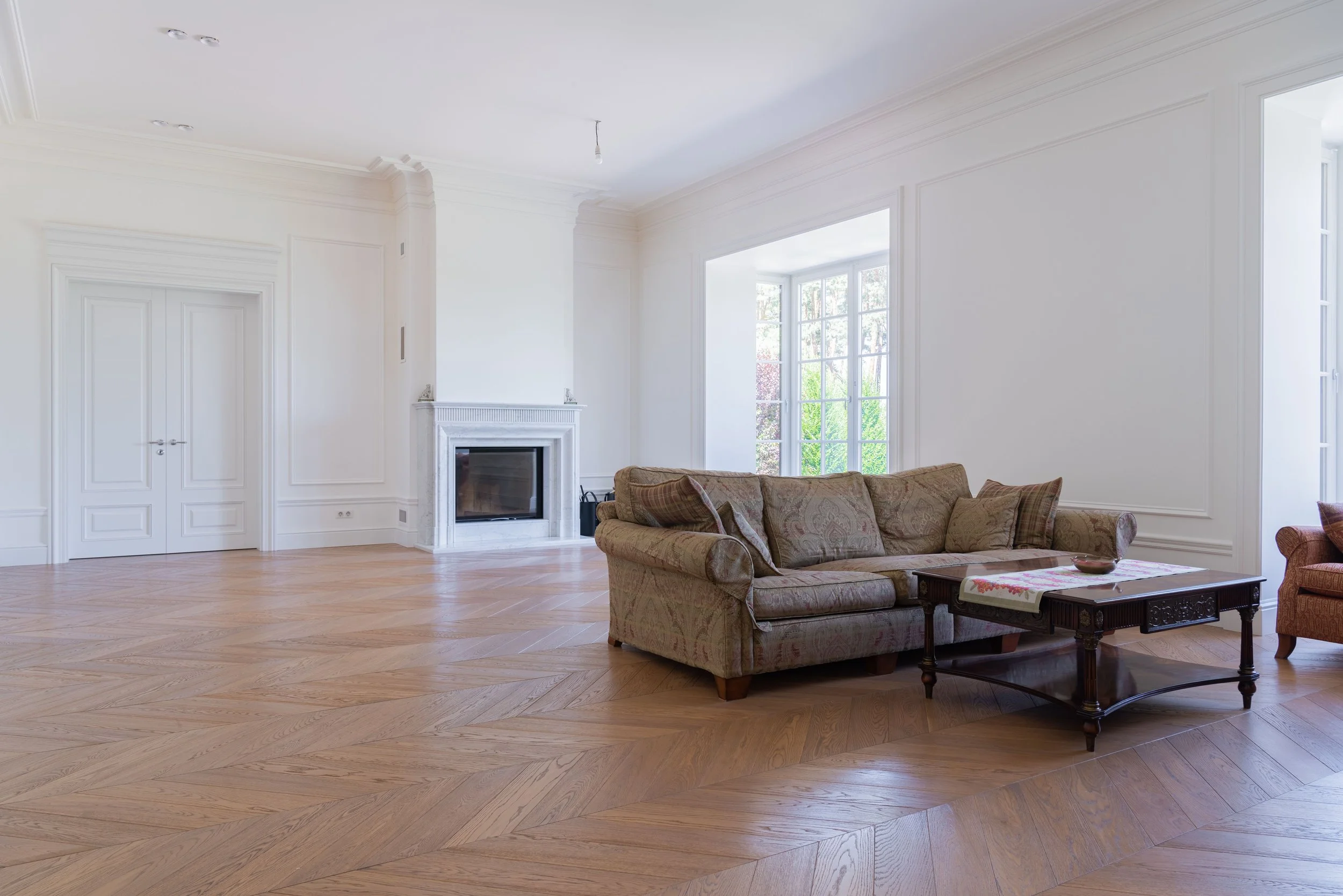 A spacious living room with white walls, a white fireplace, large windows letting in natural light, wooden flooring, and a patterned sofa with cushions. BitBrick - Cleveland's team for flooring, kitchen and bath remodels, and general construction.