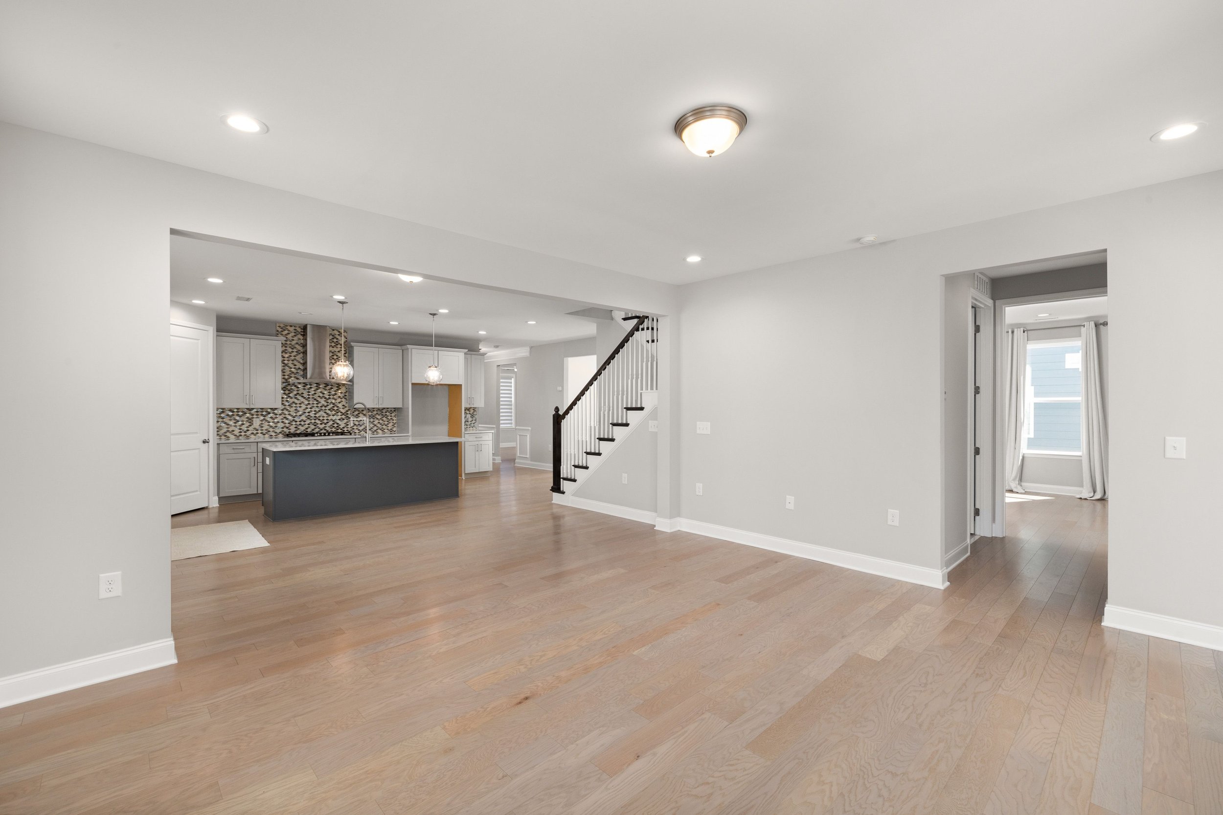 Open-concept living area with light wood flooring, white walls, kitchen with white cabinets and a mosaic backsplash, staircase with dark wood railing, and large windows with white curtains. BitBrick Cleveland Construction. Licensed & Insured.