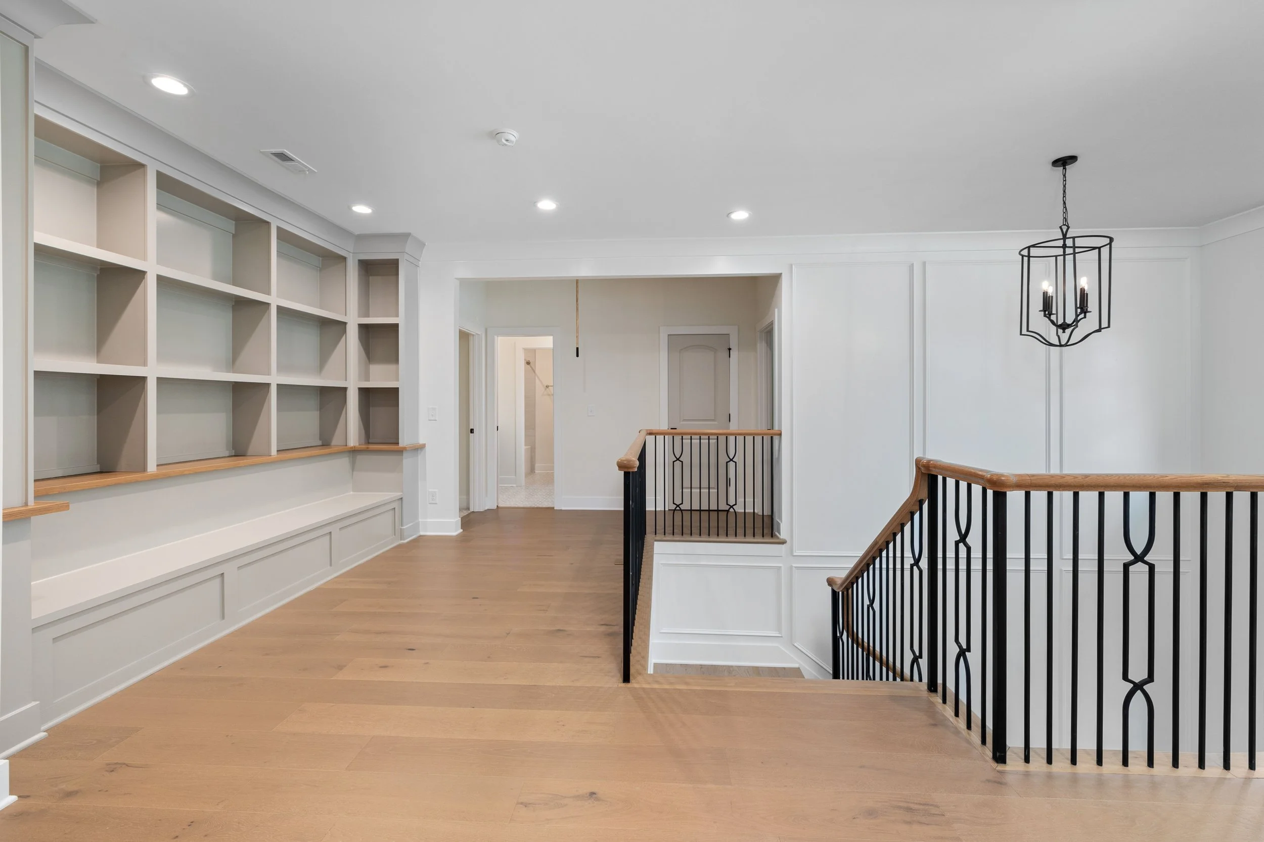 Empty room with wooden floors, white walls, built-in shelves, a hanging light fixture, and a staircase with black metal railings. BitBrick - Cleveland's local team for flooring, kitchen and bath remodels, and general construction. Licensed & Insured.