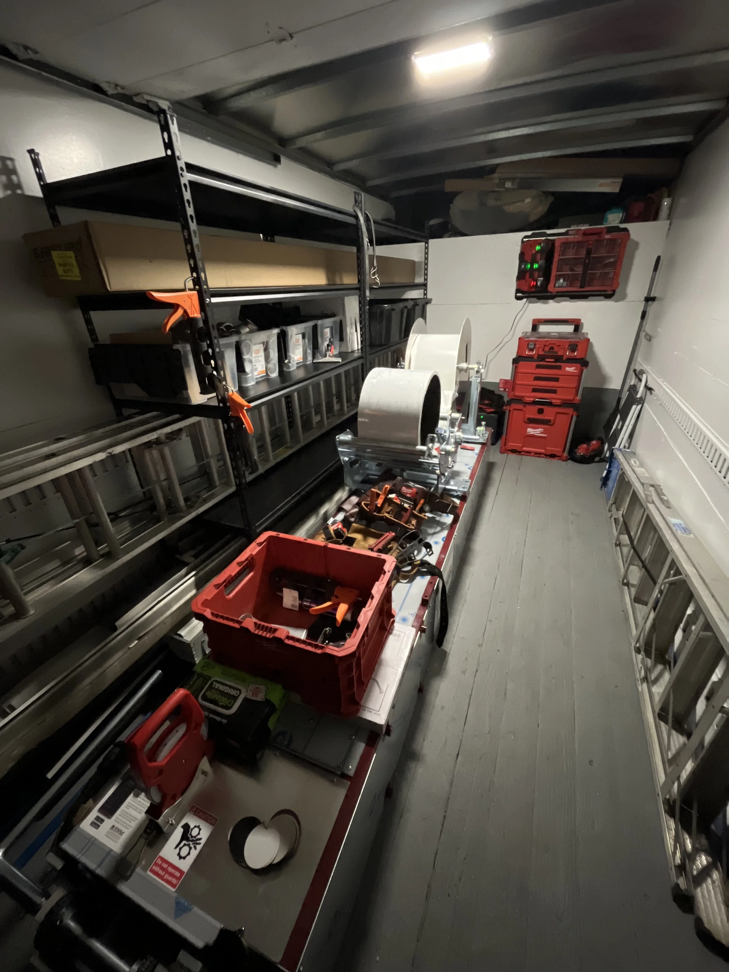 A storage room with shelves containing tools, boxes, and equipment. There are various toolboxes, a large rolls of tape, a ladder, and other hardware supplies organized in the space.