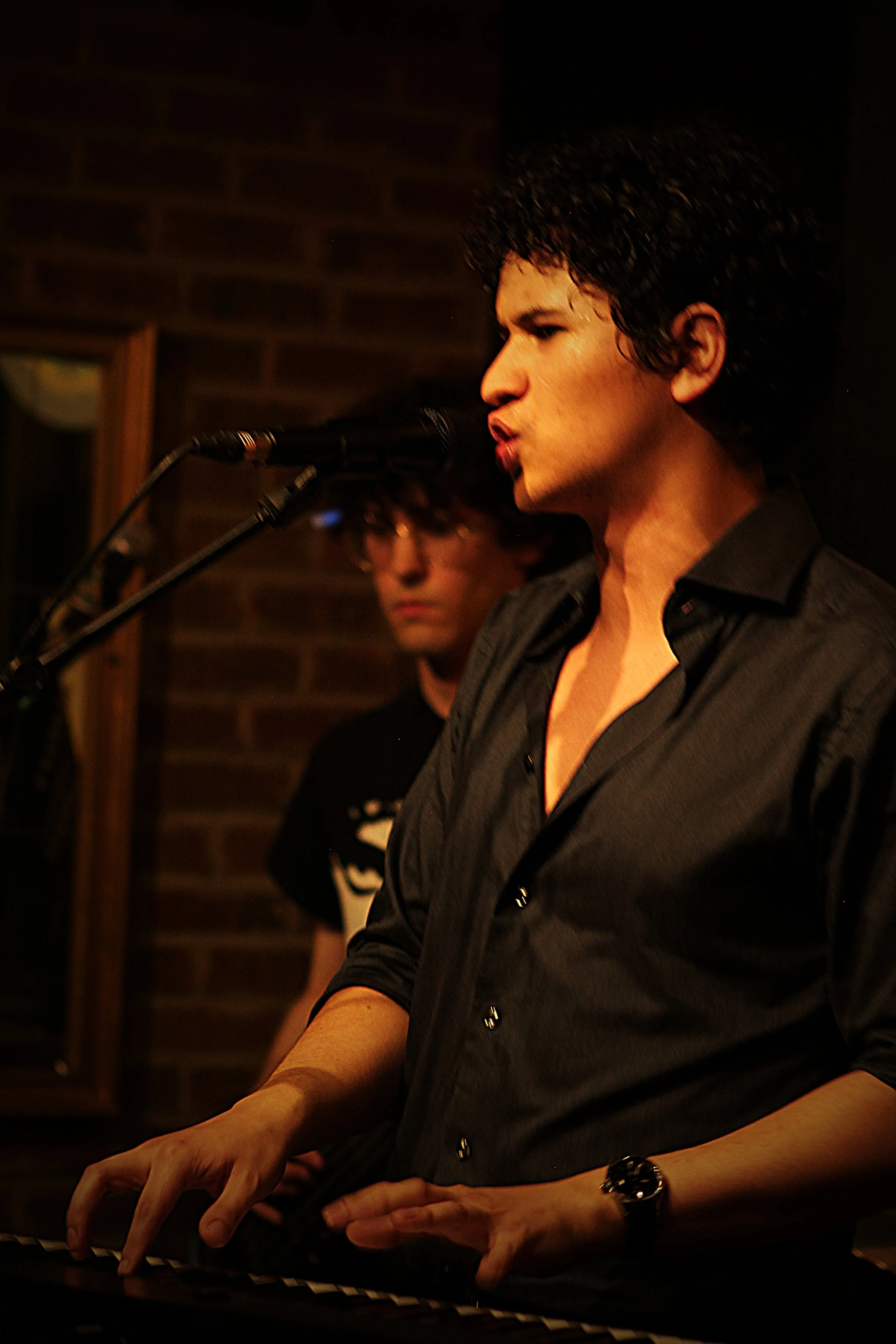 A woman with curly dark hair, wearing a black shirt, playing piano and singing into a microphone, with a man with glasses in the background against a brick wall.