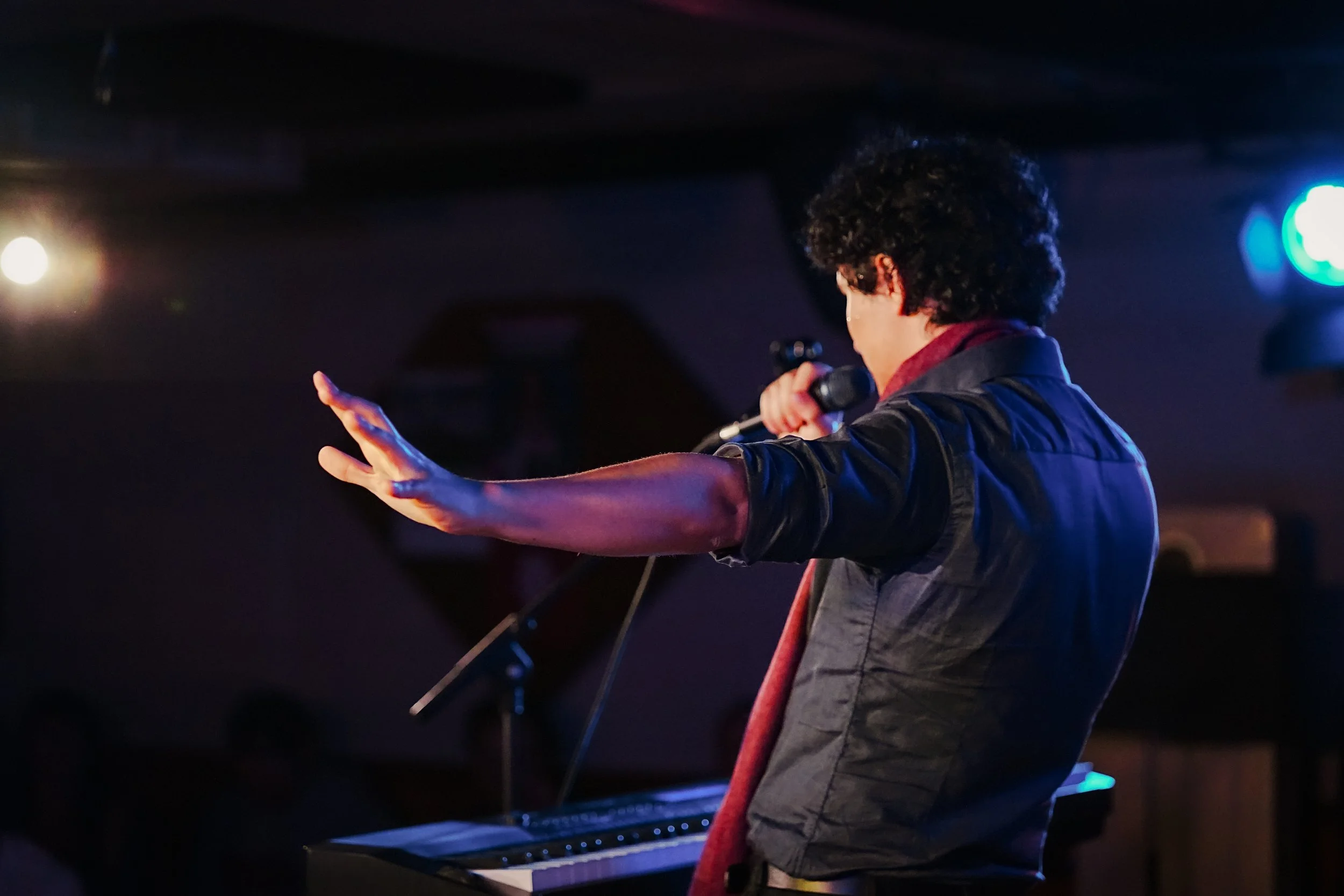 Person performing on stage with a microphone in hand, gesturing with the other hand, standing near a keyboard with stage lights in the background.
