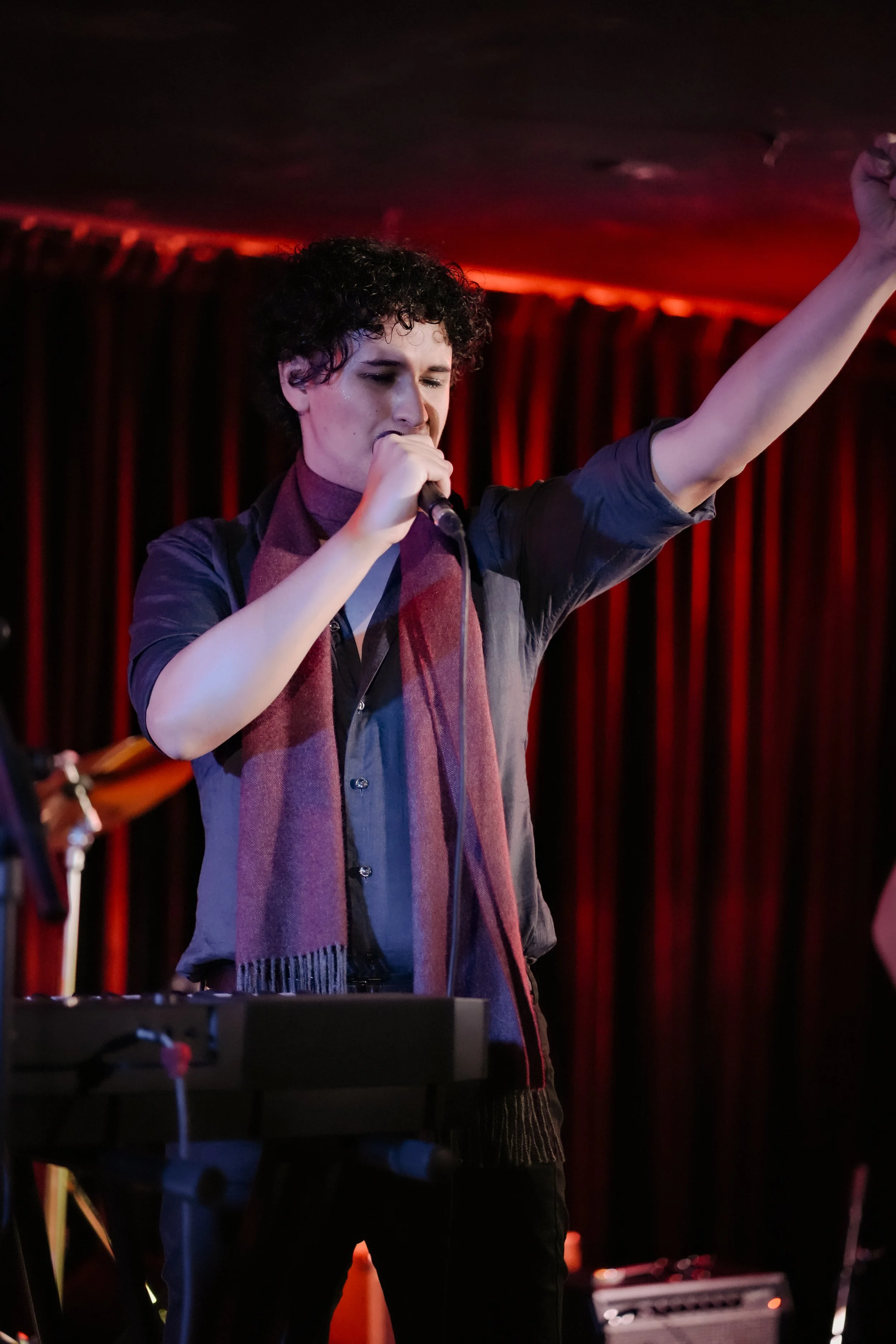 A person with curly black hair singing into a microphone with their eyes closed, raising one arm in a performance on stage with red curtains, music equipment, and a keyboard in the foreground.