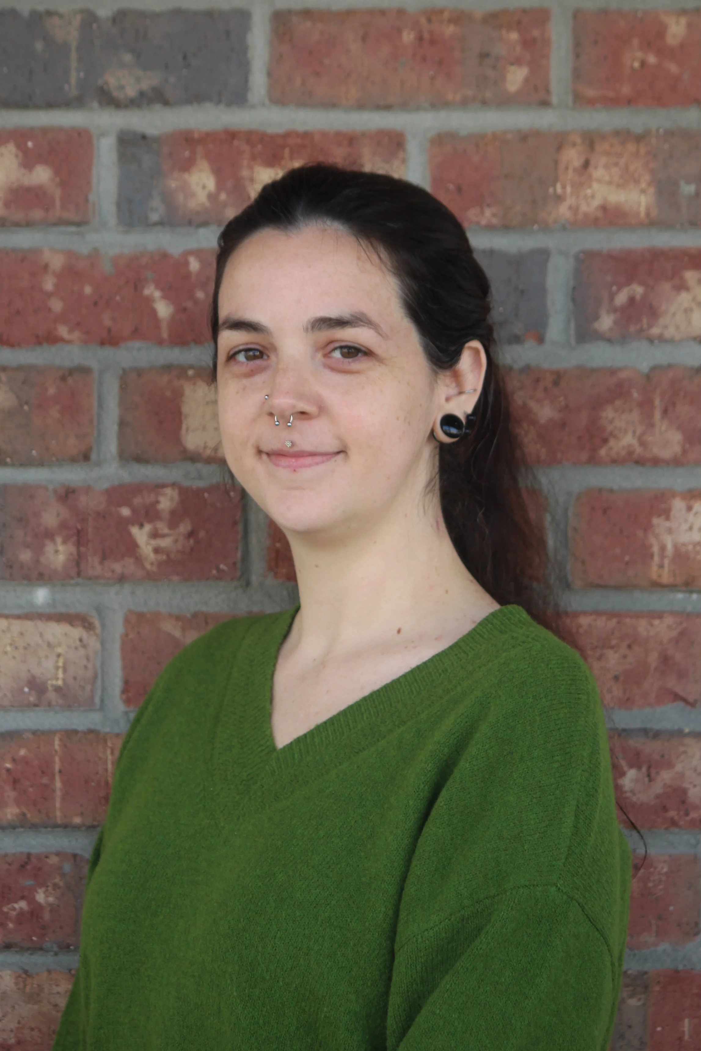 A woman with dark hair pulled back, light skin, and multiple facial piercings, wearing a green sweater, standing against a red brick wall.