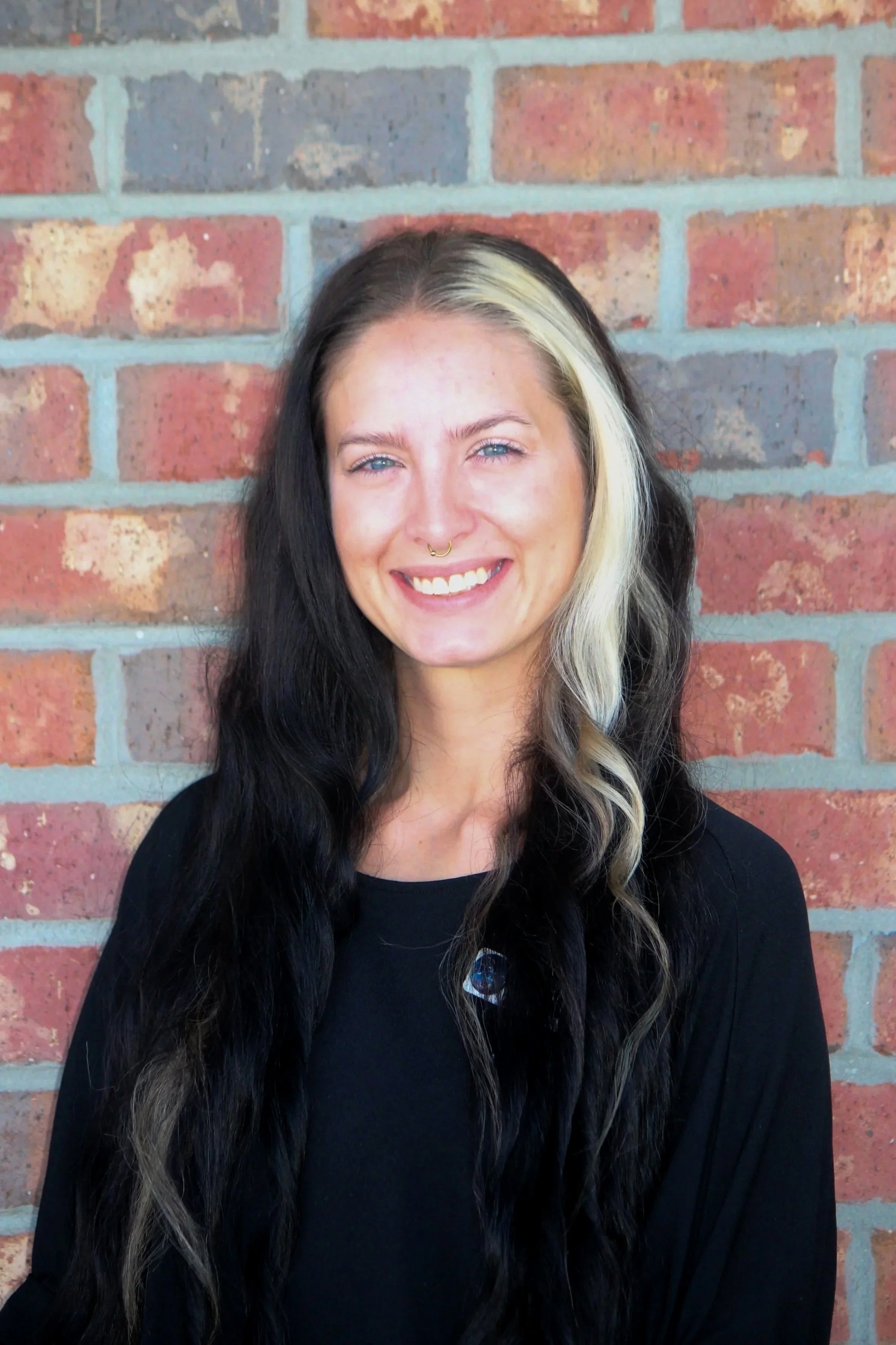 A woman with black and blonde hair smiling in front of a brick wall.