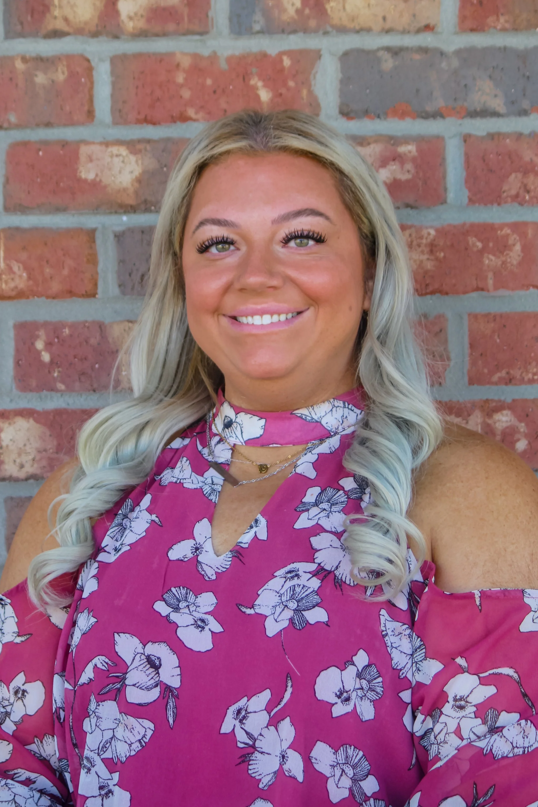 A woman with long blonde hair, wearing a pink floral off-shoulder top and necklaces, standing in front of a brick wall, smiling at the camera.