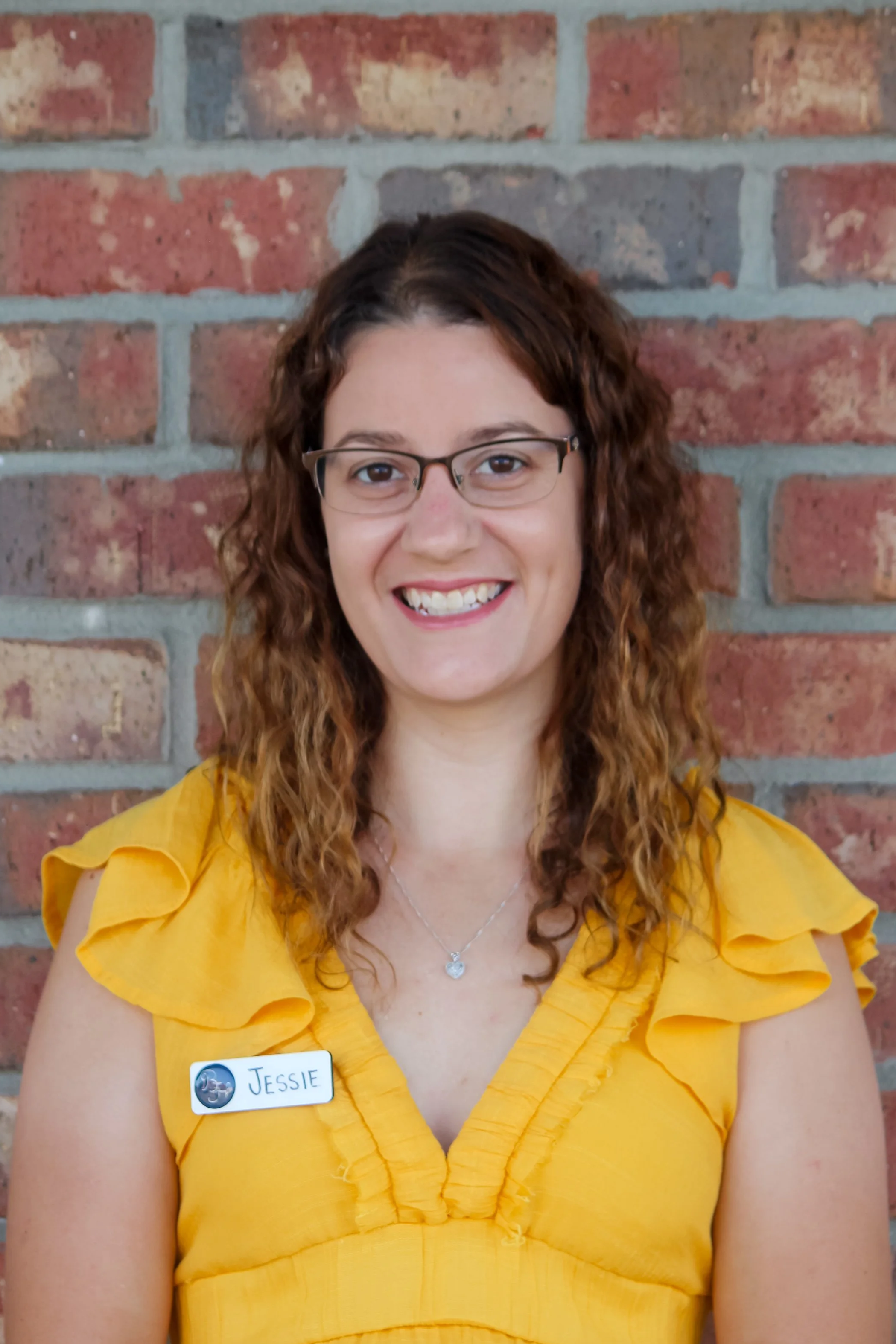 Woman with curly hair and glasses smiling, wearing a yellow ruffled blouse, standing against a brick wall. She has a name tag that reads 'Jessie'.