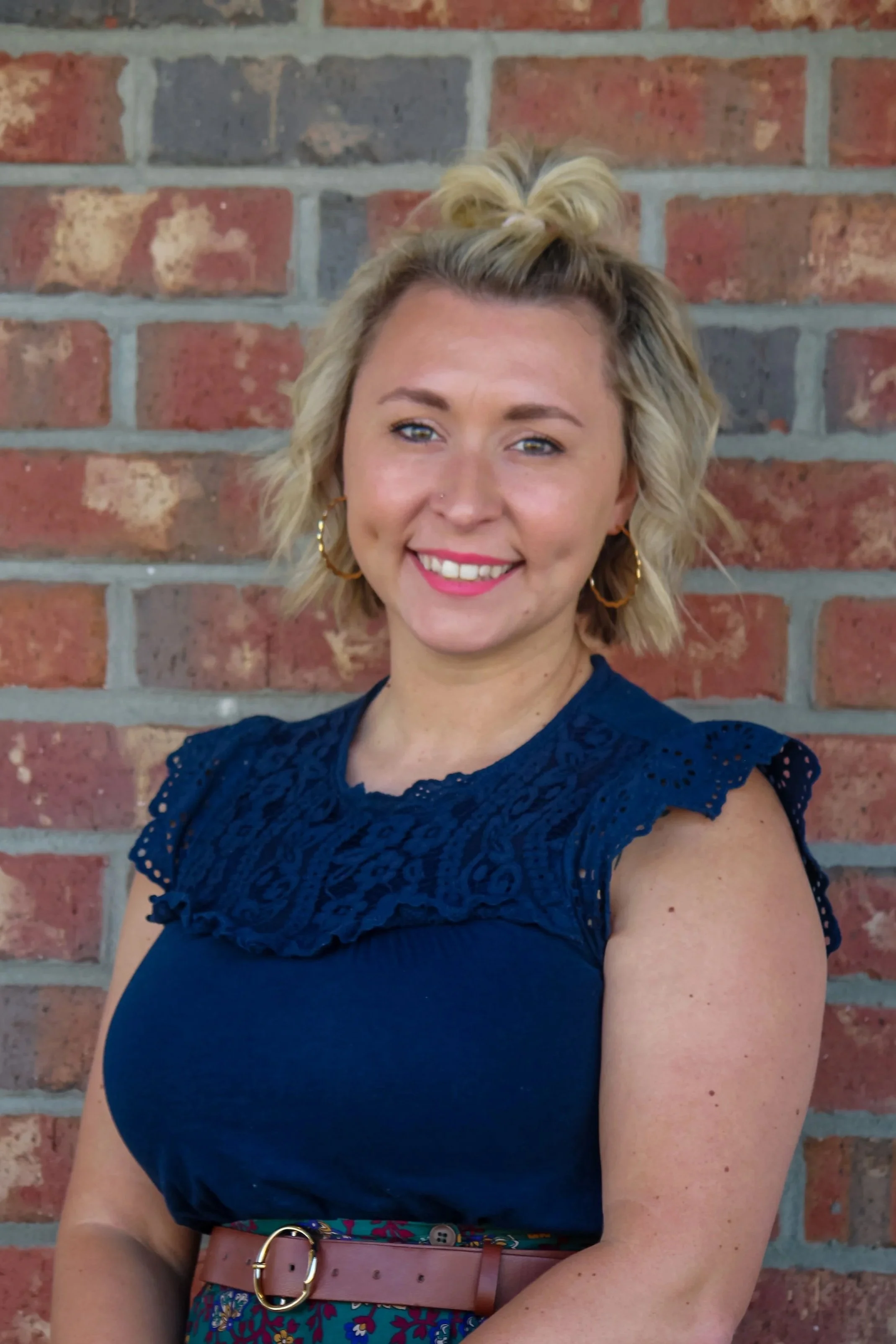 A woman with blonde hair styled in a half-up top knot, wearing hoop earrings and a navy blue top with lace details, standing in front of a brick wall.
