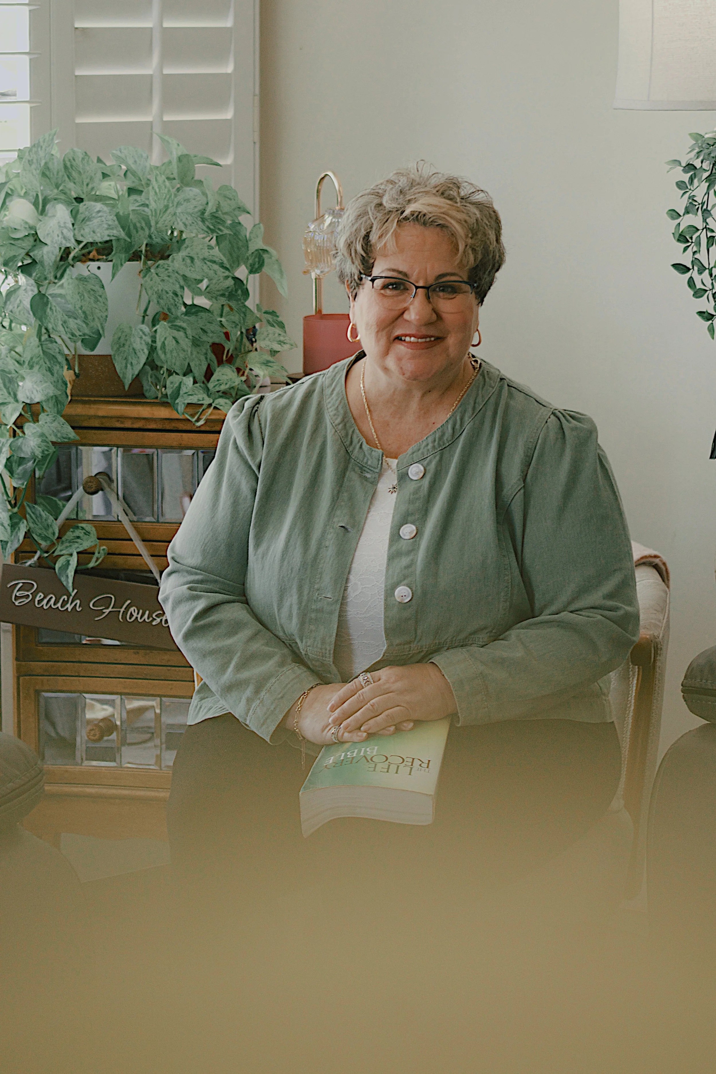 Bess Leto, a middle-aged woman with short curly hair, glasses, and gold jewelry, sitting at a table with a book titled 'LIFE RECOVERY' in front of her, in a room with plants and a wooden cabinet.