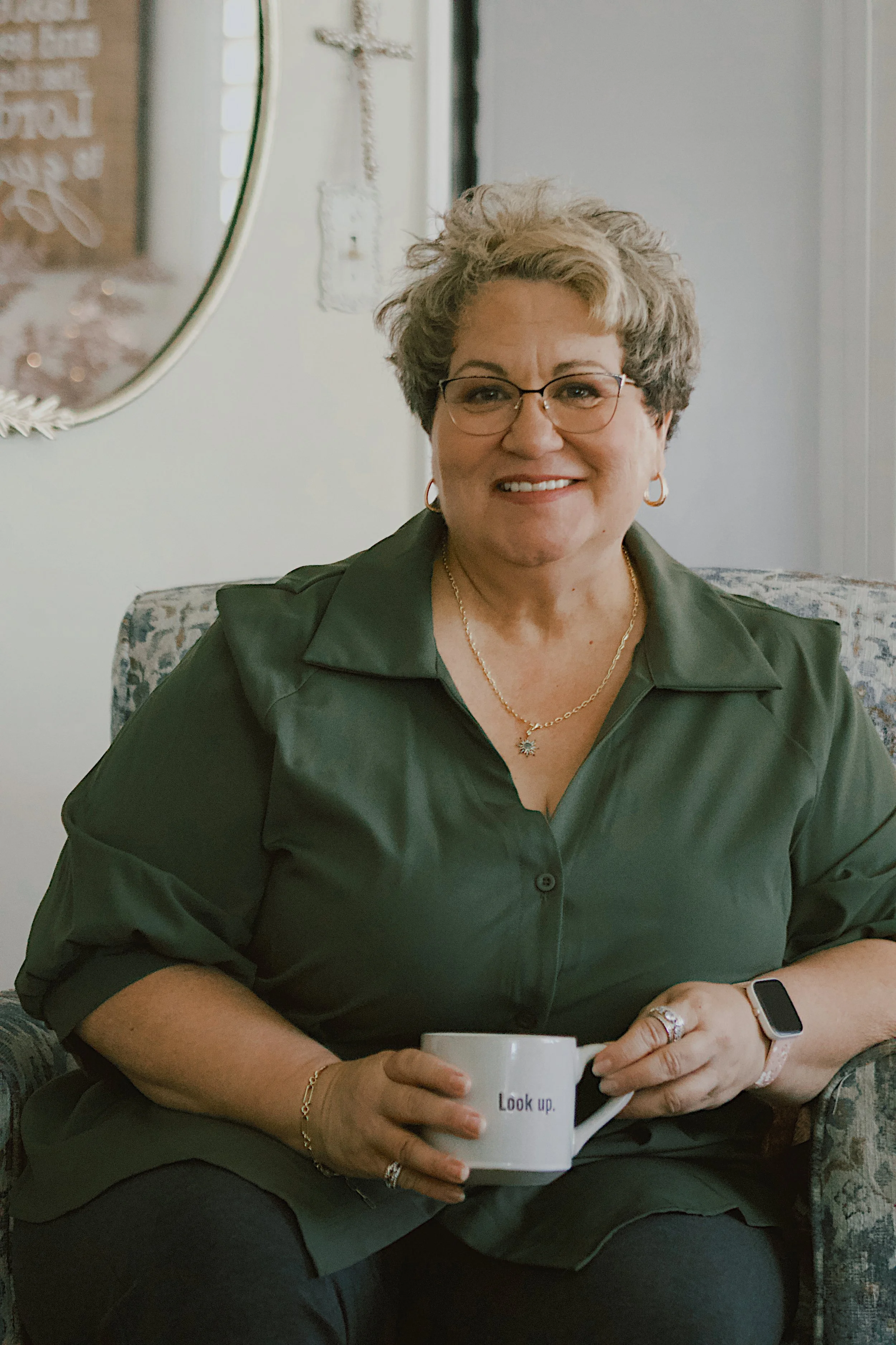 Bess Leto, a woman with short gray hair, glasses, and jewelry, sitting indoors at a table, holding a white coffee mug with the text 'Look up.' Bess is is smiling, and there is a mirror and a decorative cross on the wall in the background.
