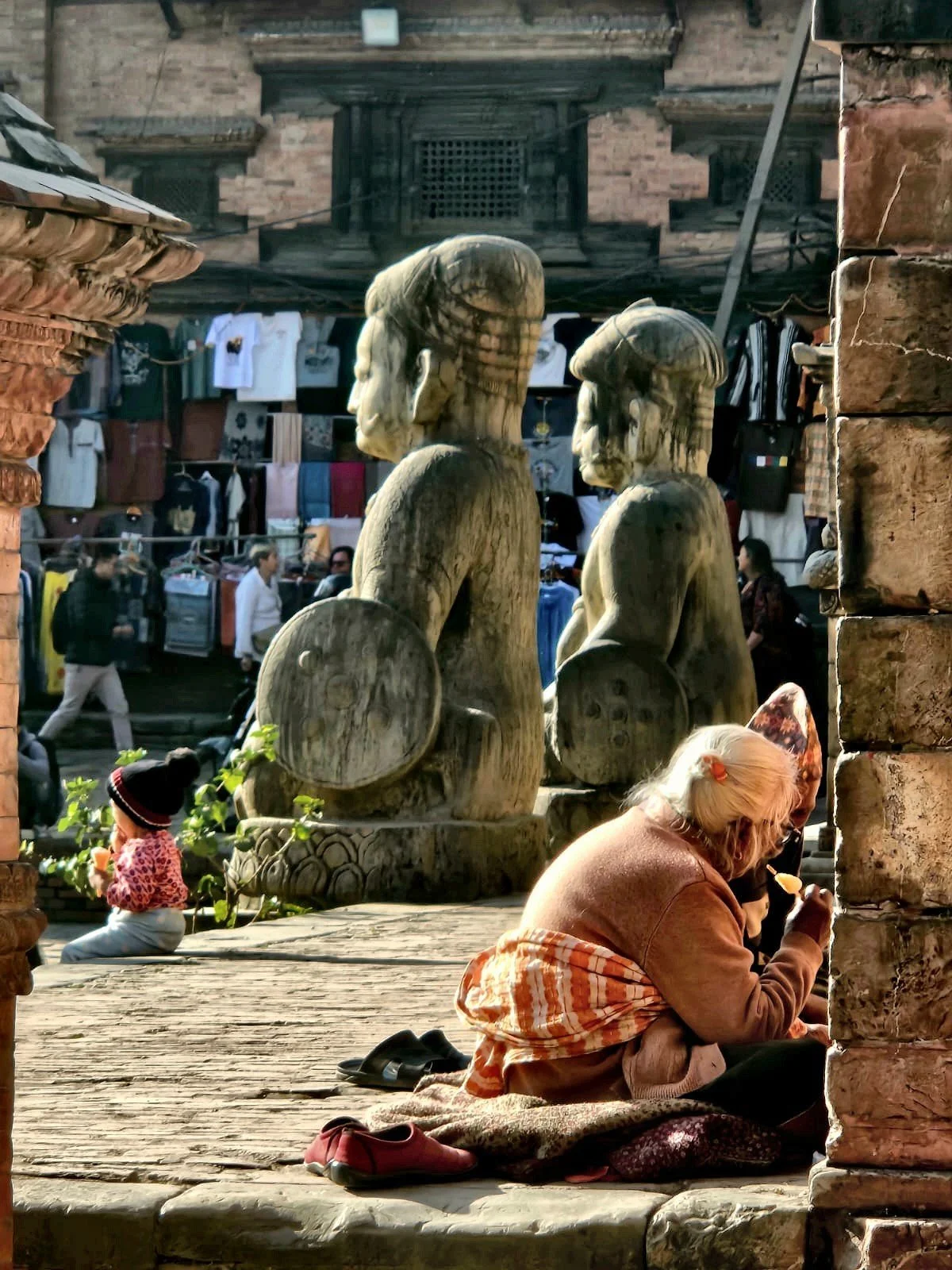 Donna seduta accanto a statue di elefanti in pietra, in un affollato mercato storico. Baktapur