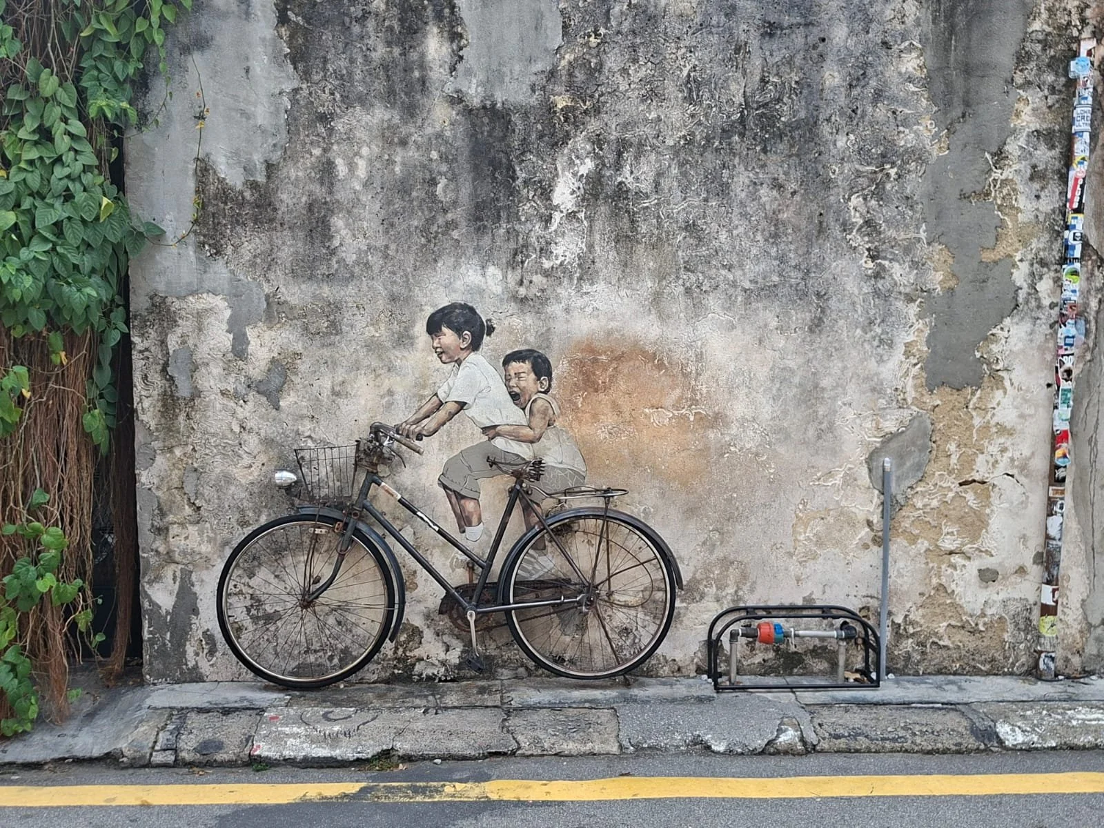 Murale urbano raffigurante una donna in bicicletta con un bambino su una strada acciottolata, su un vecchio muro ricoperto di graffiti e vegetazione. Malesia George Town