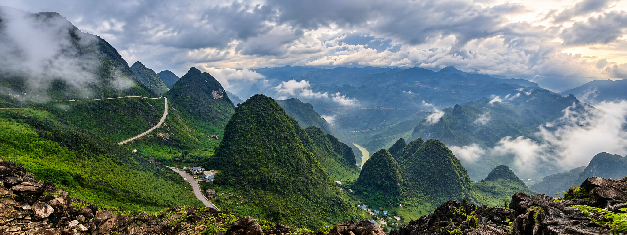Panorama di montagne verdi e cime carsiche avvolte dalla nebbia, con strada tortuosa, villaggi e fiume nella valle. Vietnam