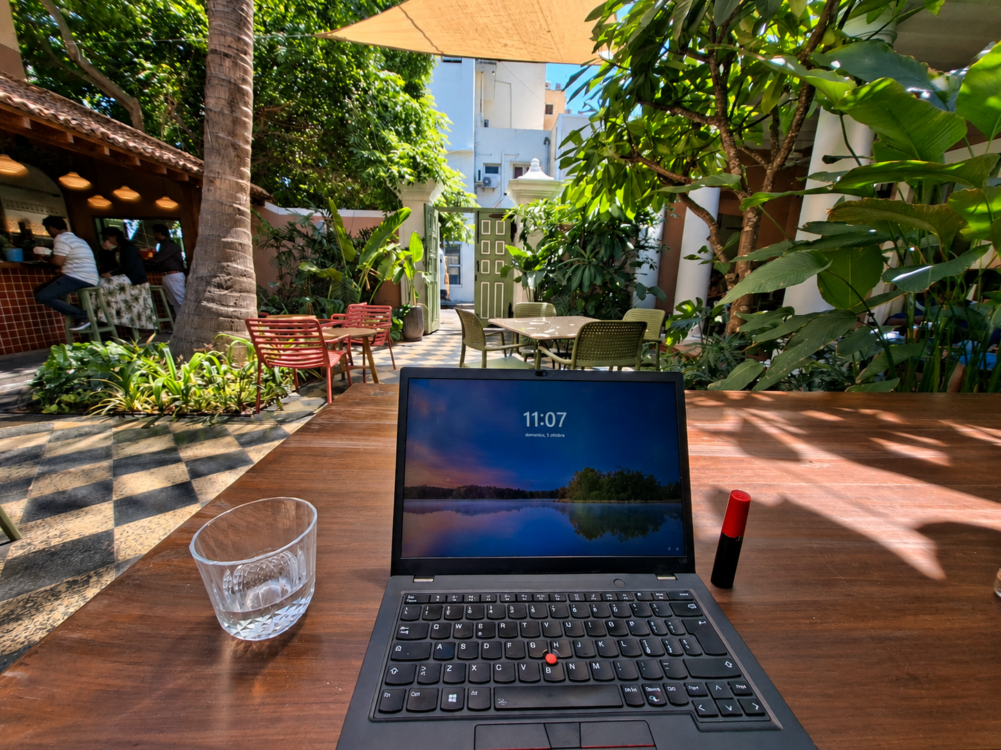 Laptop su un tavolo in legno in un cortile tropicale di un caffè, con piante, palme e tavoli all’aperto. Pondicherry
