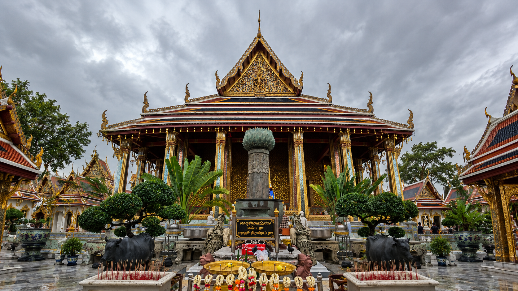 Tempio buddista thailandese riccamente decorato con tetti dorati, colonne colorate, statue di bufali e offerte votive sotto un cielo nuvoloso.