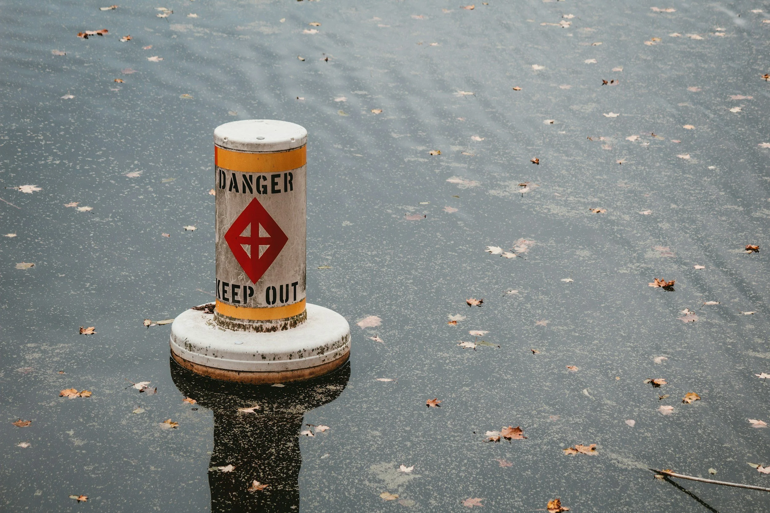 A white cylindrical danger warning pole with orange and black markings, standing in a body of water covered with fallen leaves and small debris.