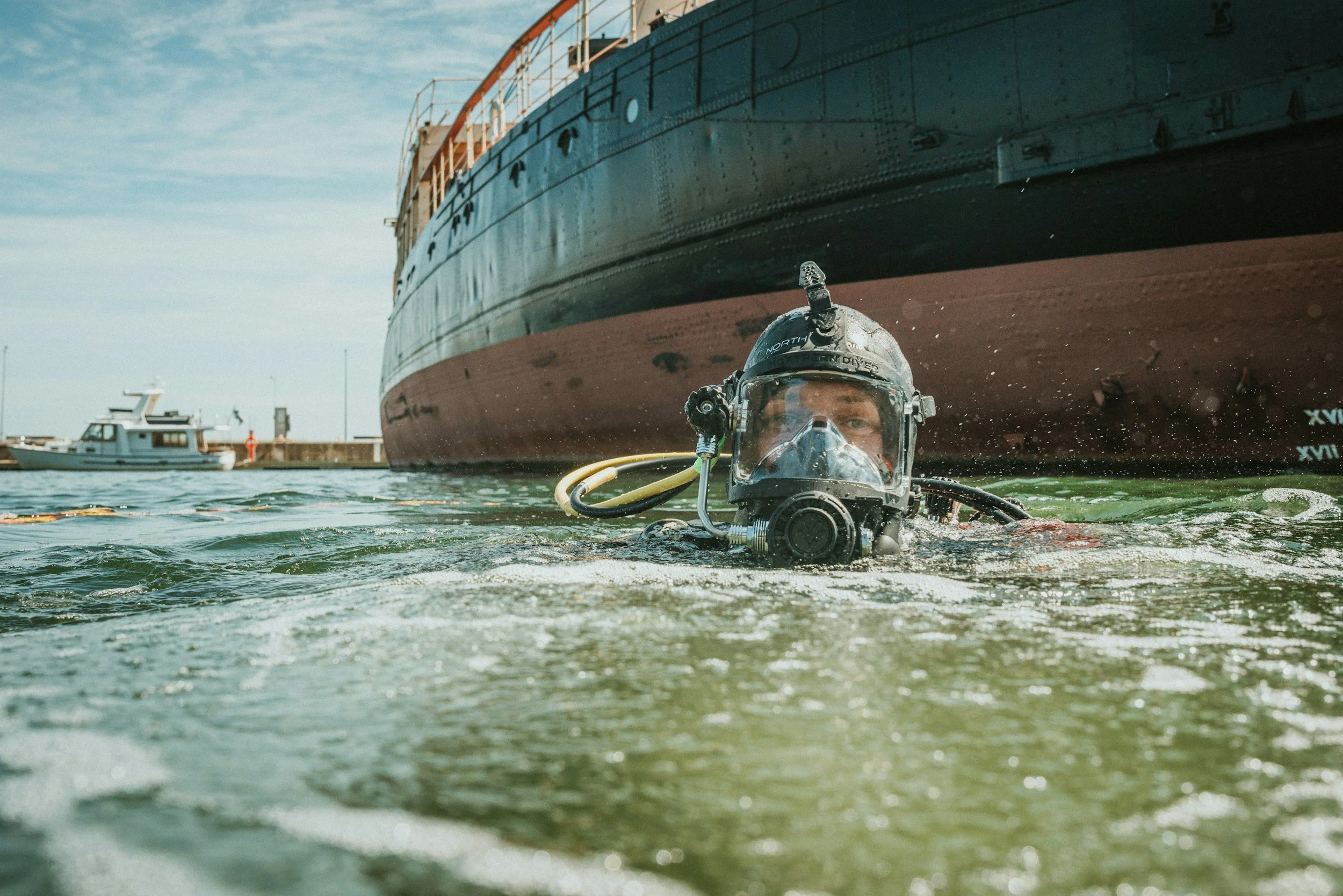 A diver wearing a helmet and diving gear submerged in water near a large ship in a harbor, with smaller boats visible in the background.