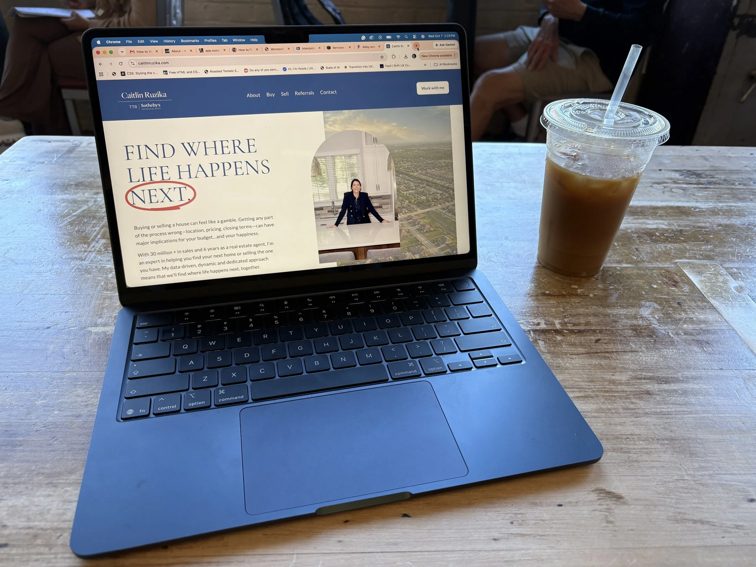 A laptop on a wooden table displaying a real estate website, next to a plastic cup filled with iced coffee with a straw.