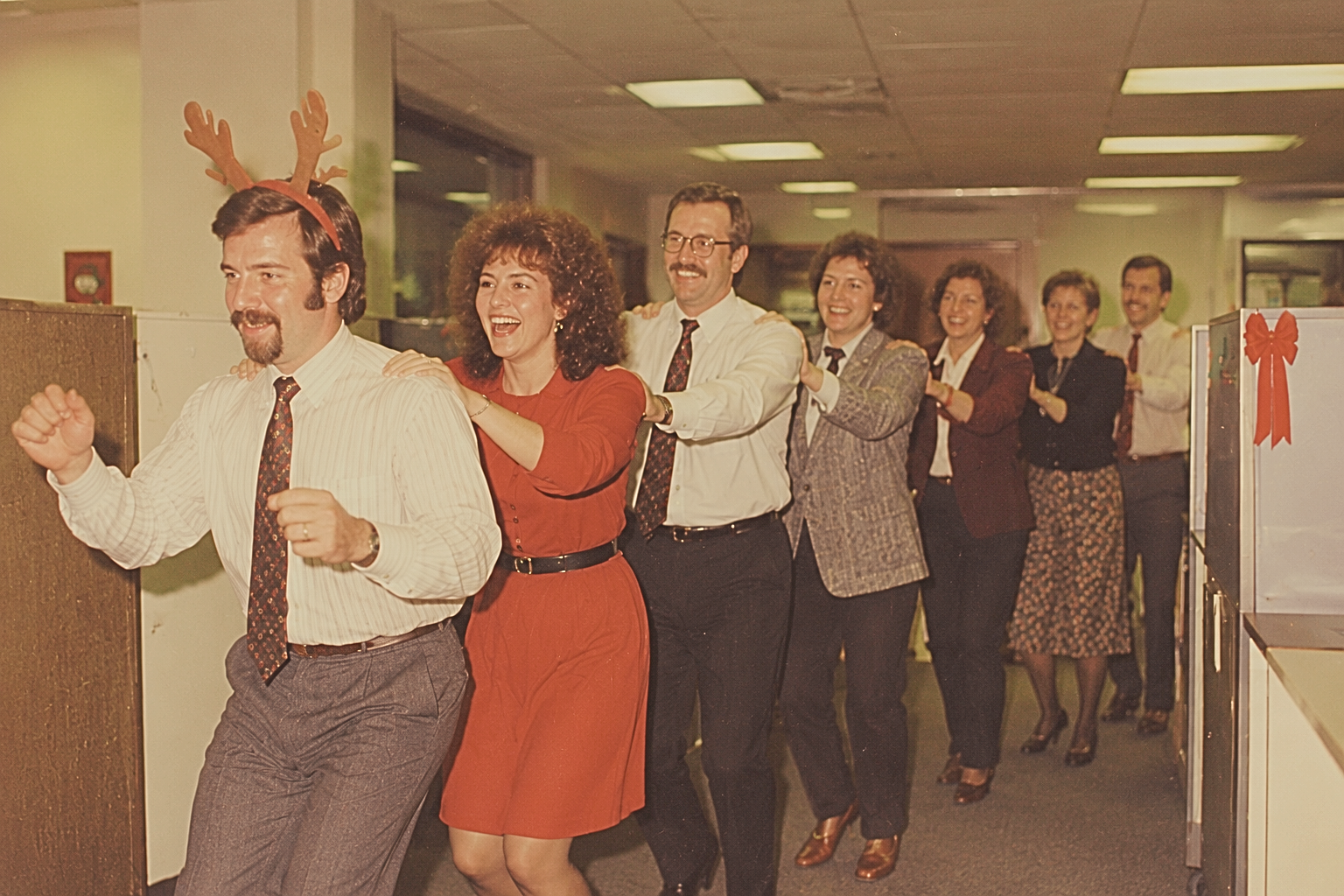 Office workers in holiday attire doing a conga line, led by a man wearing reindeer antlers.