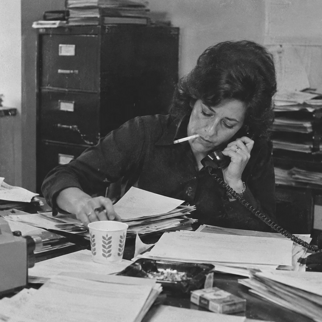 Black and white photo of a woman working at a cluttered desk, talking on a phone, with a cigarette in her mouth, surrounded by papers and office supplies.