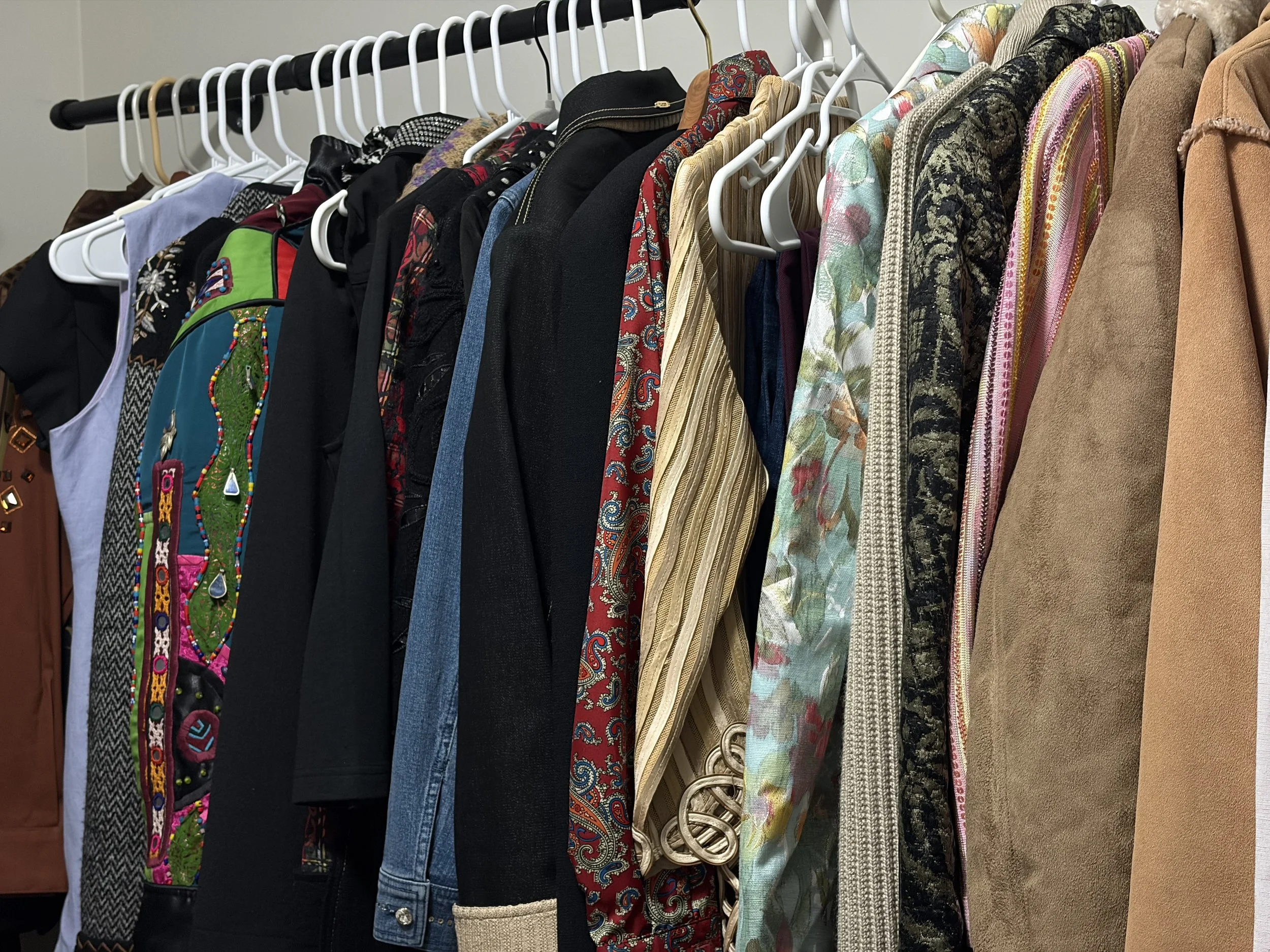 Clothing rack filled with various jackets, suits, and tops in different colors and patterns, including floral, striped, and solid fabrics.