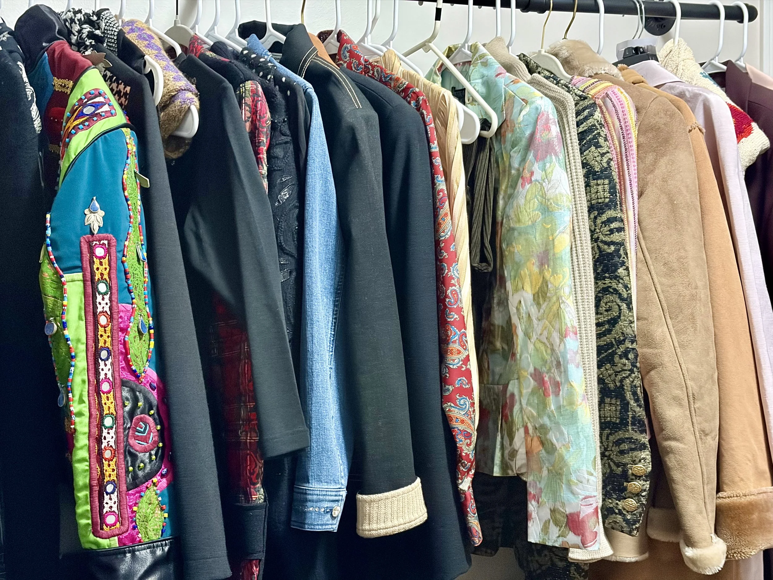 Clothes hanging on a rack including jackets, shirts, dresses, and sweaters in various colors and patterns.