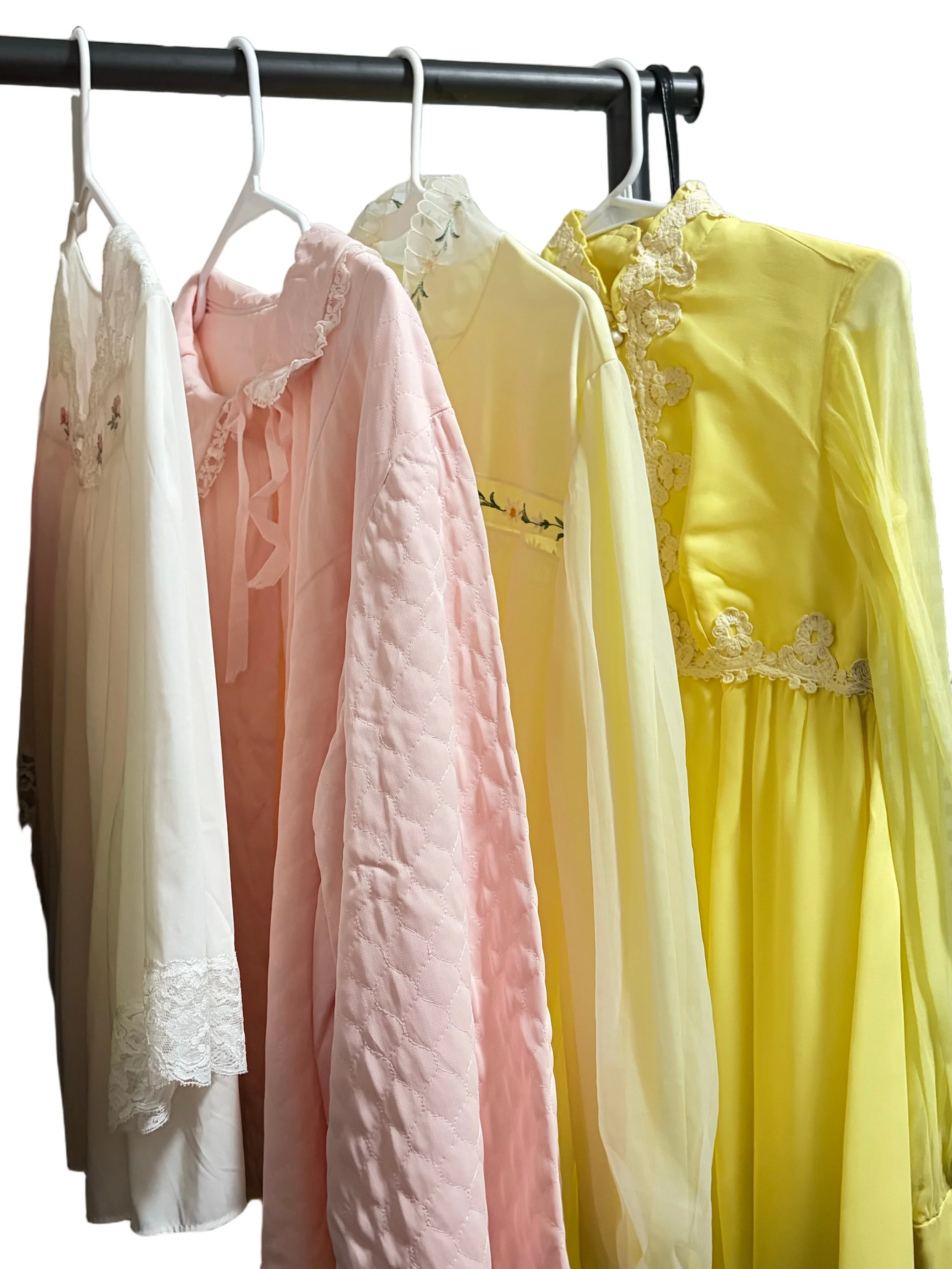 A row of colorful dresses hanging on a clothing rack. The dresses are in shades of white, pink, yellow, and have various lace and embroidery details.