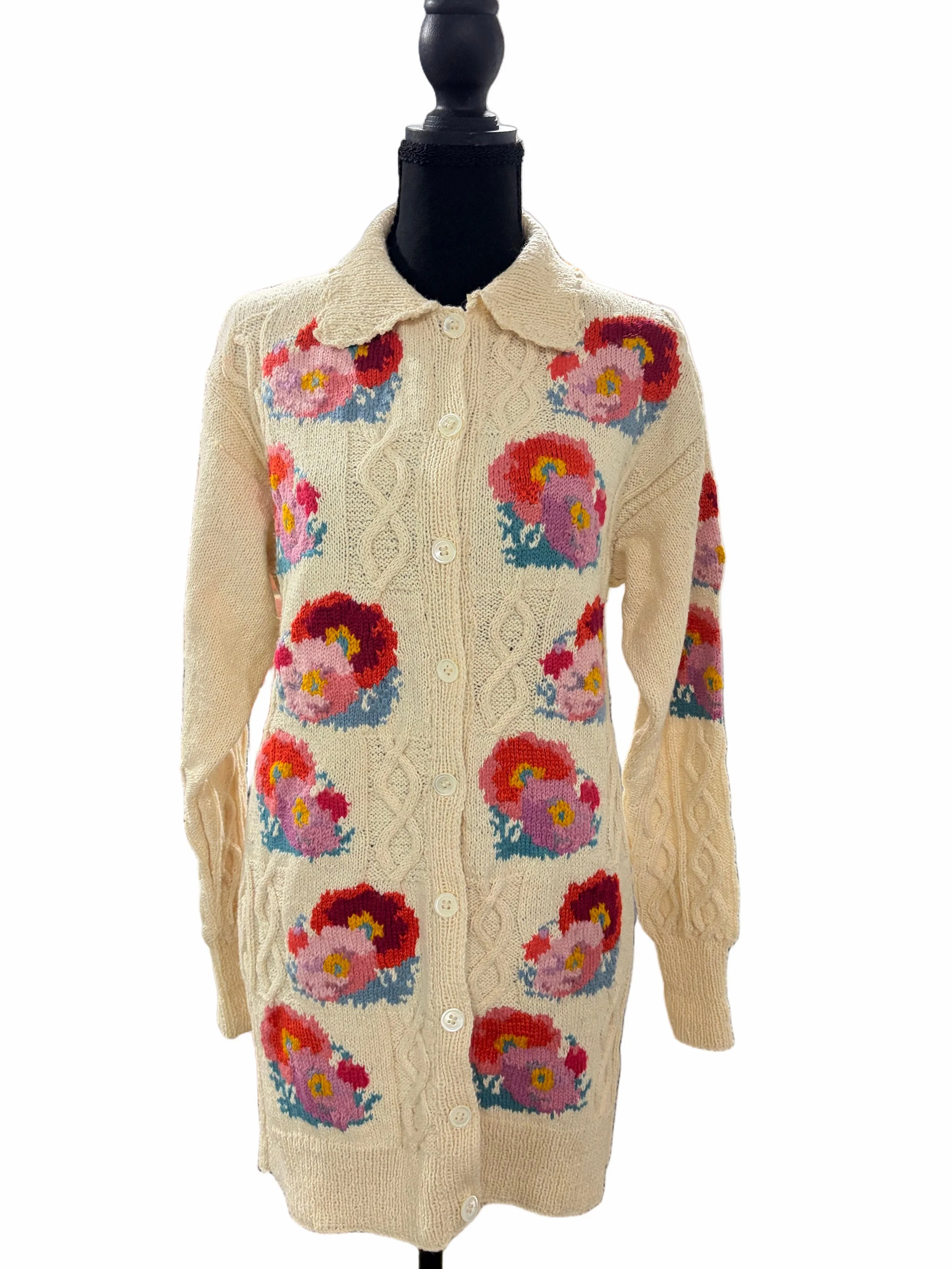 A cream-colored knitted cardigan with colorful floral patterns, buttons down the front, and a collar on a black mannequin.