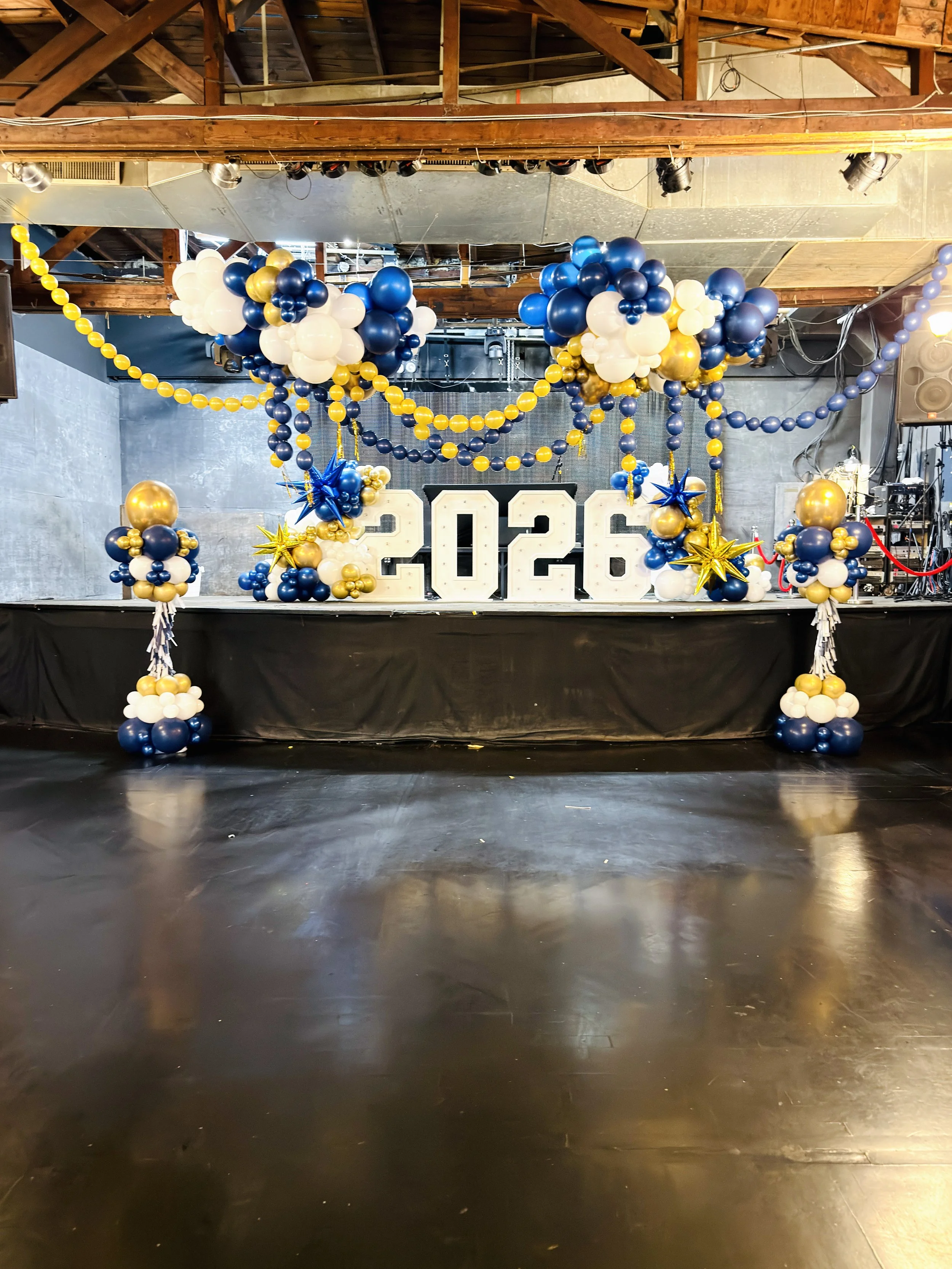 Stage decorated with blue, white, and gold balloons, large number cutouts displaying 2026, and star-shaped balloons for a New Year celebration.