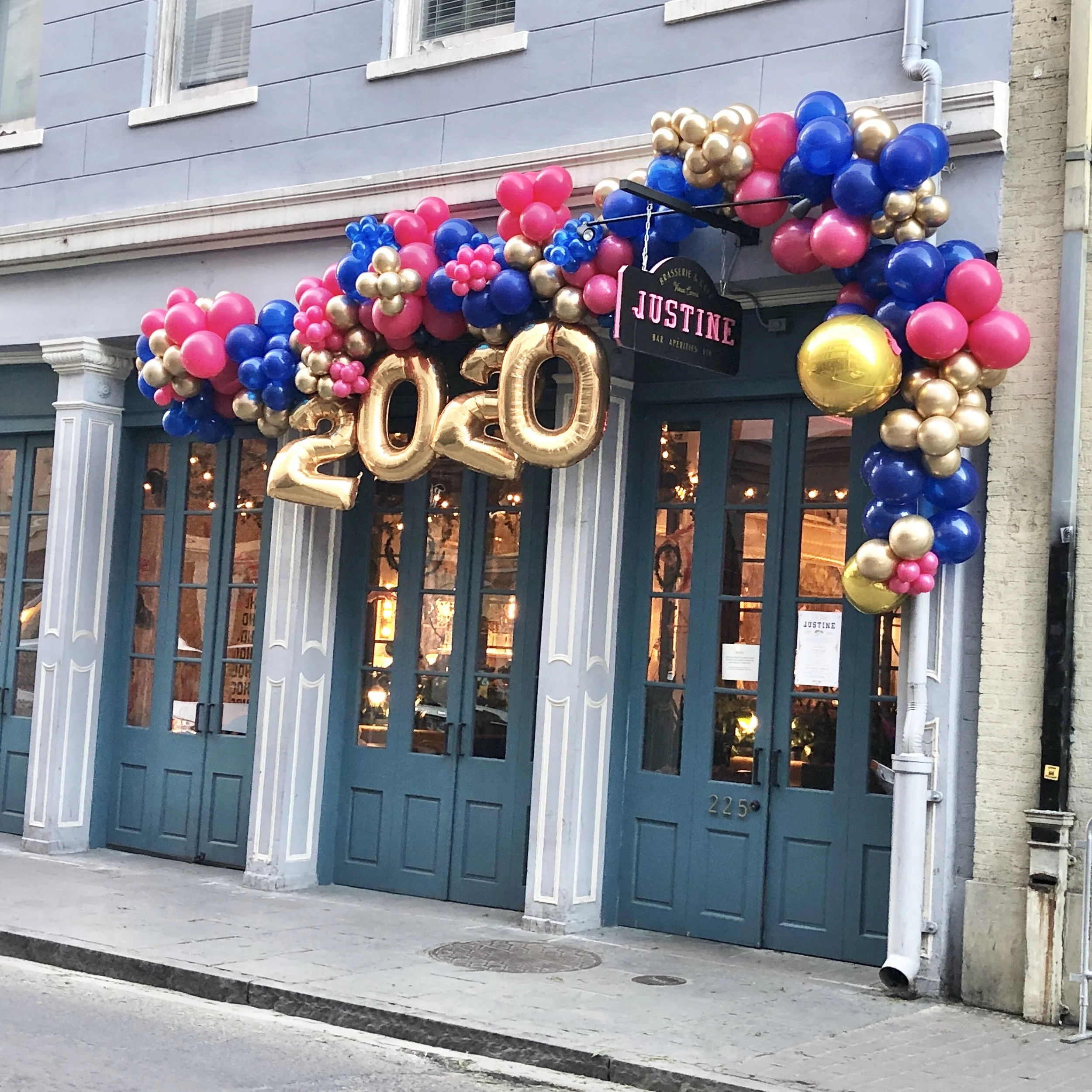 Storefront decorated with pink, blue, and gold balloons, including gold foil number balloons forming '2023', and a hanging sign for Justine bar and cuisine, on a city street.