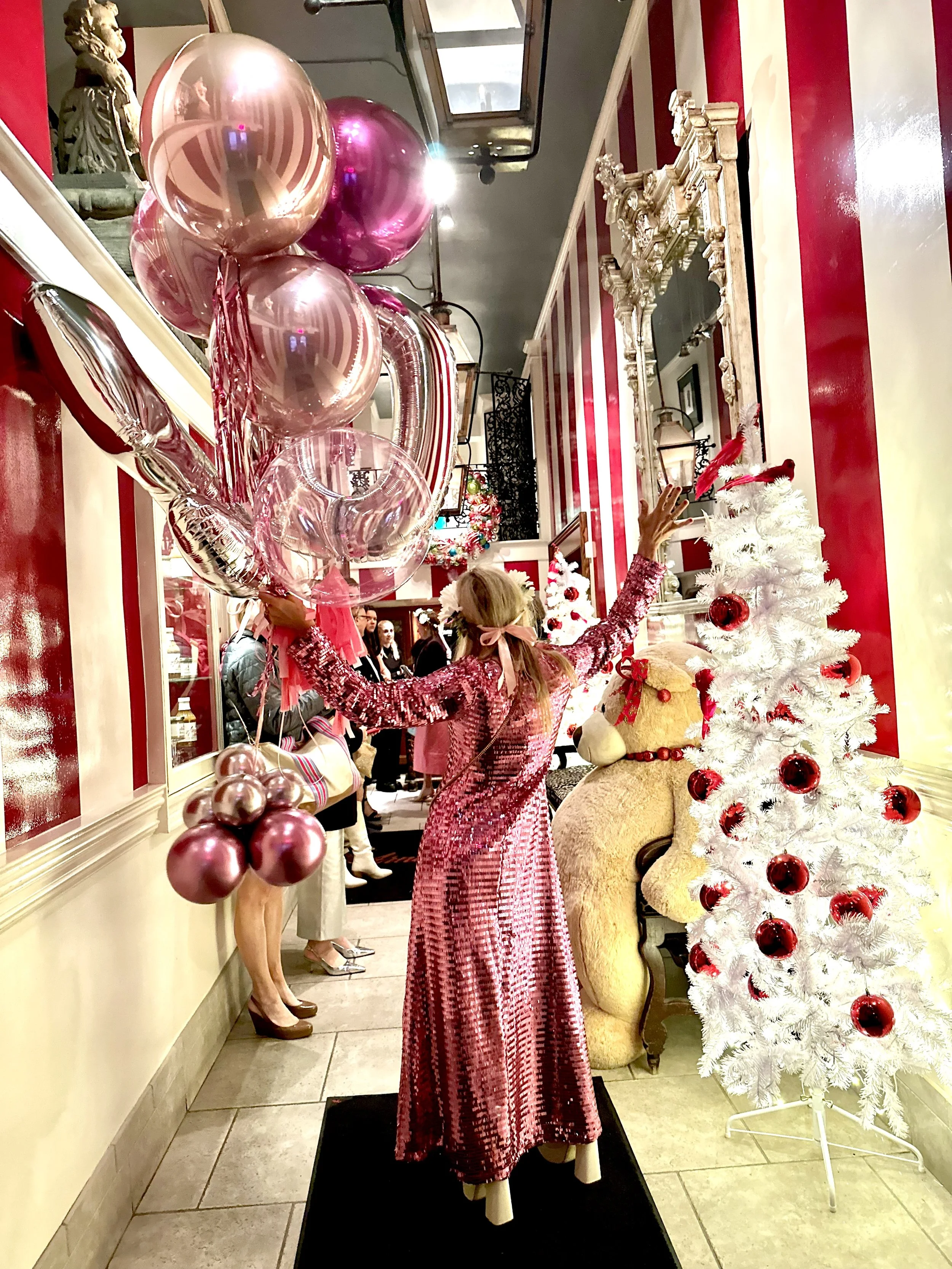 A store window display with a mannequin dressed in a red patterned dress, surrounded by Christmas decorations including white and red Christmas trees, a bear stuffed animal, and festive ornaments. The display is decorated for Christmas with balloons, ribbons, and holiday-themed decor.
