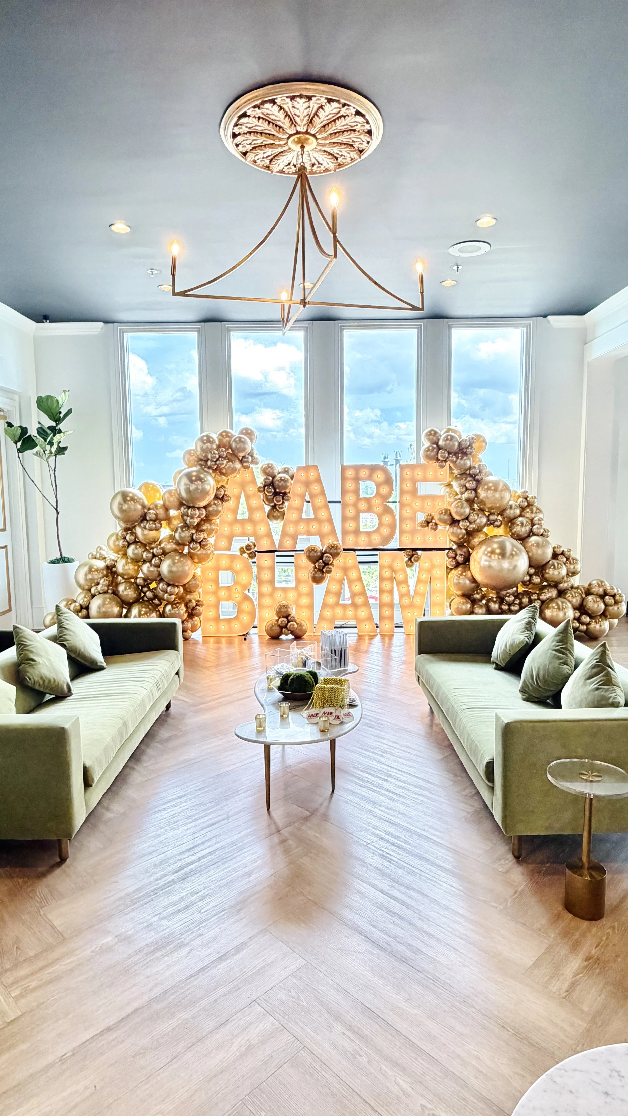 Brightly lit room with a neon sign that says 'BABE BHAM' surrounded by gold, silver, and champagne-colored balloons. Two light-colored sofas with cushions face each other, and a small table with decorations and candles is in the center. Large windows with a view of blue sky and clouds in the background.