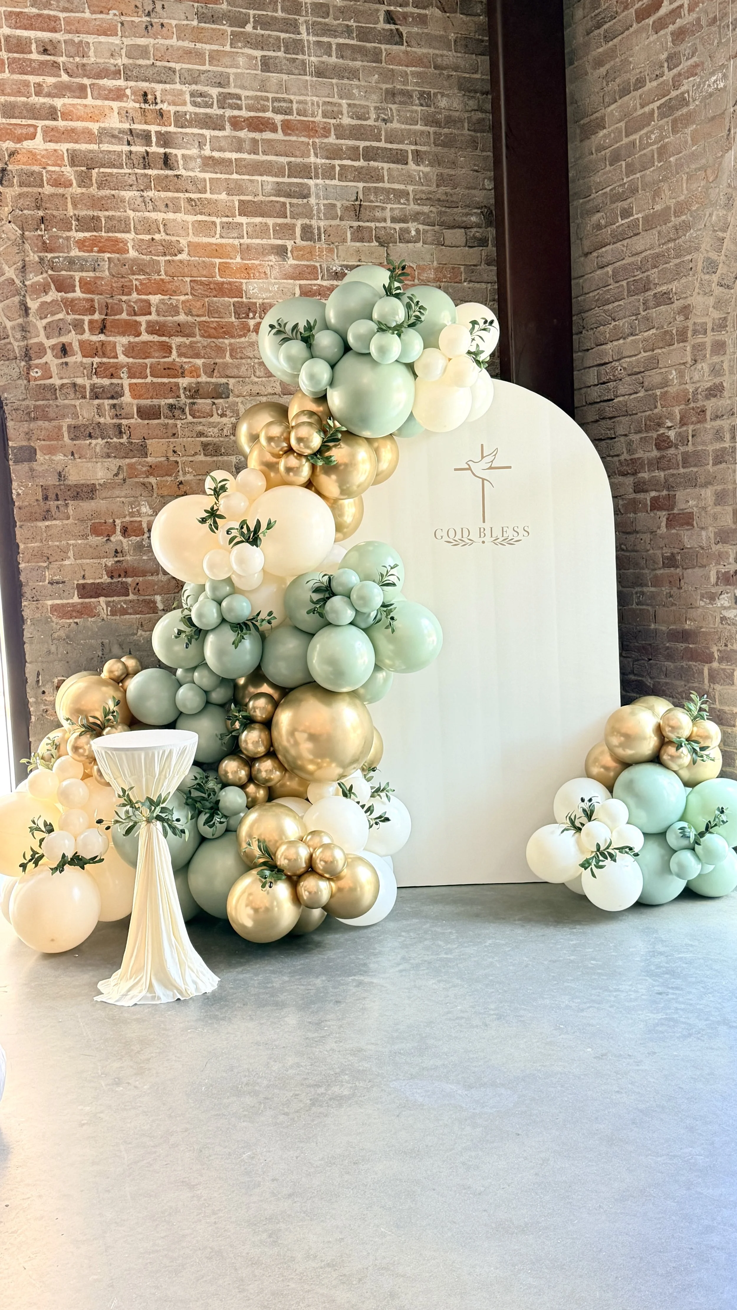Balloon arrangement with pastel gold, mint green, and white balloons, decorated with green leaves, surrounding a white backdrop with a gold cross and the words 'God Bless' written on it. A white pedestal with a pleated cloth is in front of the display, against an exposed brick wall.