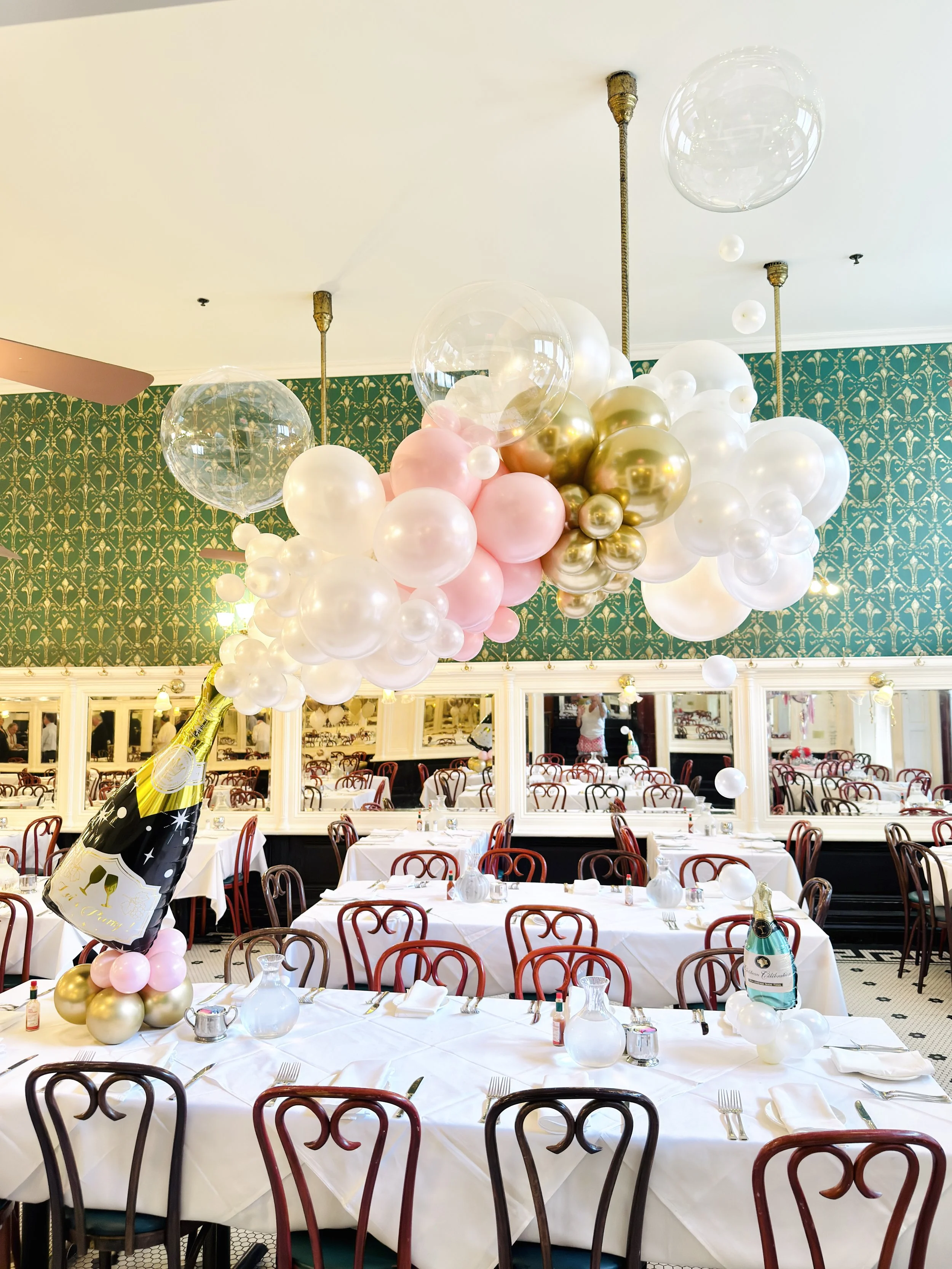 Balloon centerpiece with pink, gold, and clear balloons over a dining room table set for a celebration at a restaurant.