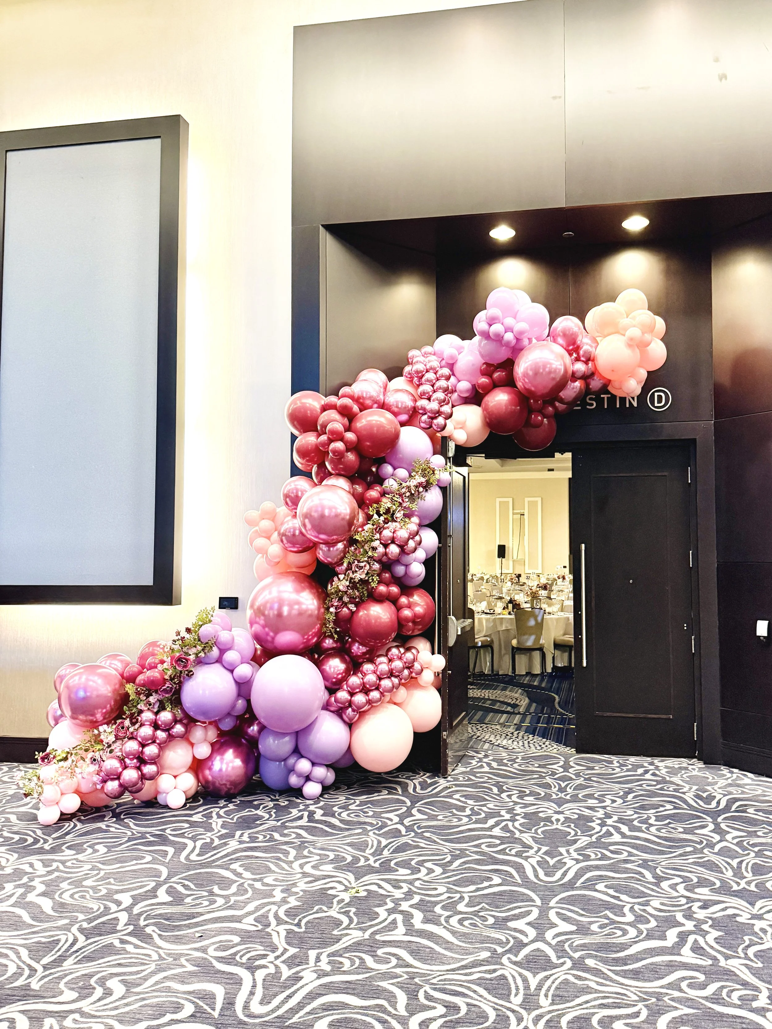 Decorated entrance with pink and purple balloons arranged in a cluster, leading into a banquet hall with tables set for an event.