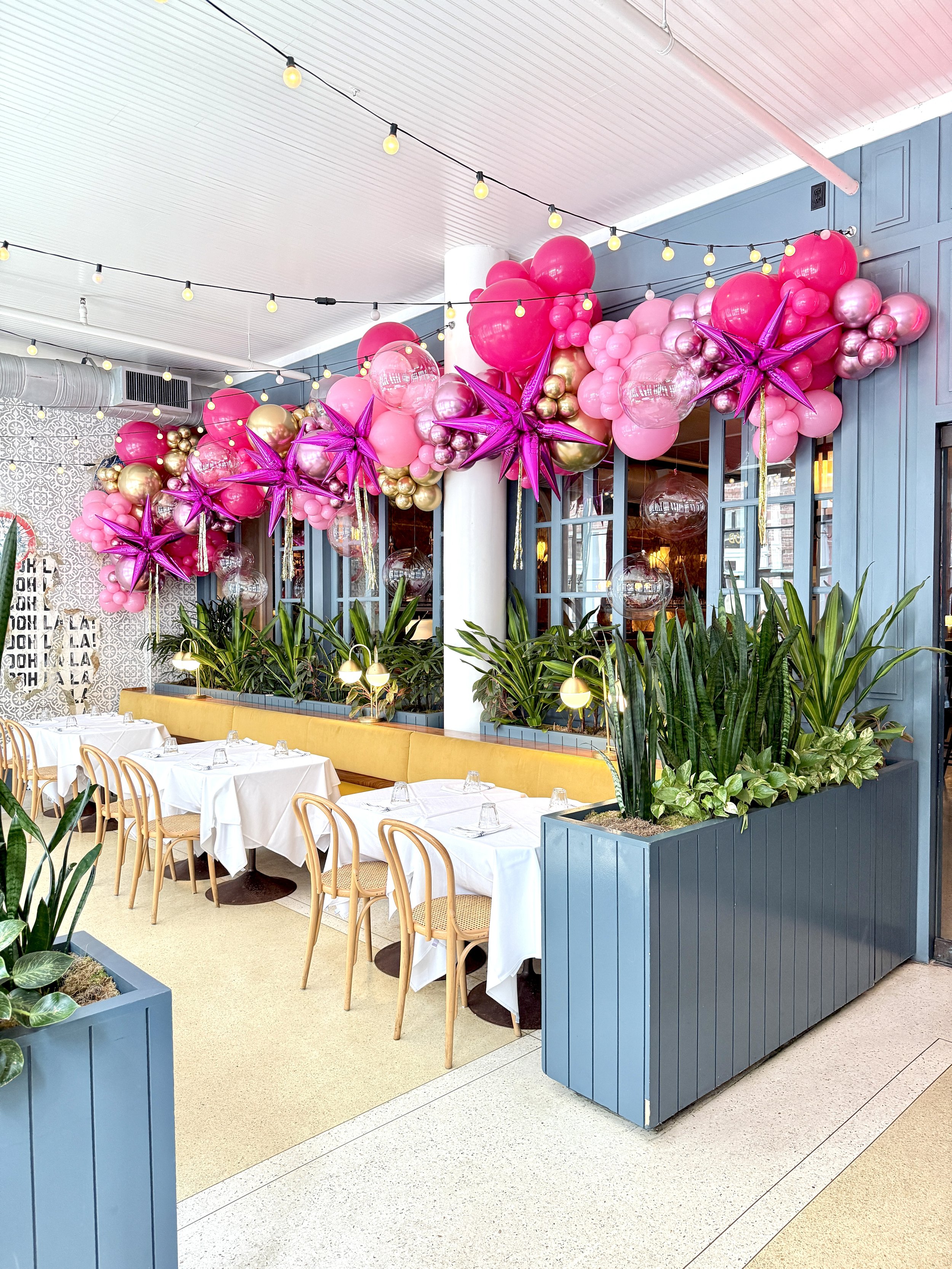 Interior of a restaurant with white tables and wooden chairs, decorated for a celebration with pink and gold balloons and purple star-shaped balloons hanging from the ceiling. There are green plants in blue planters and string lights overhead.