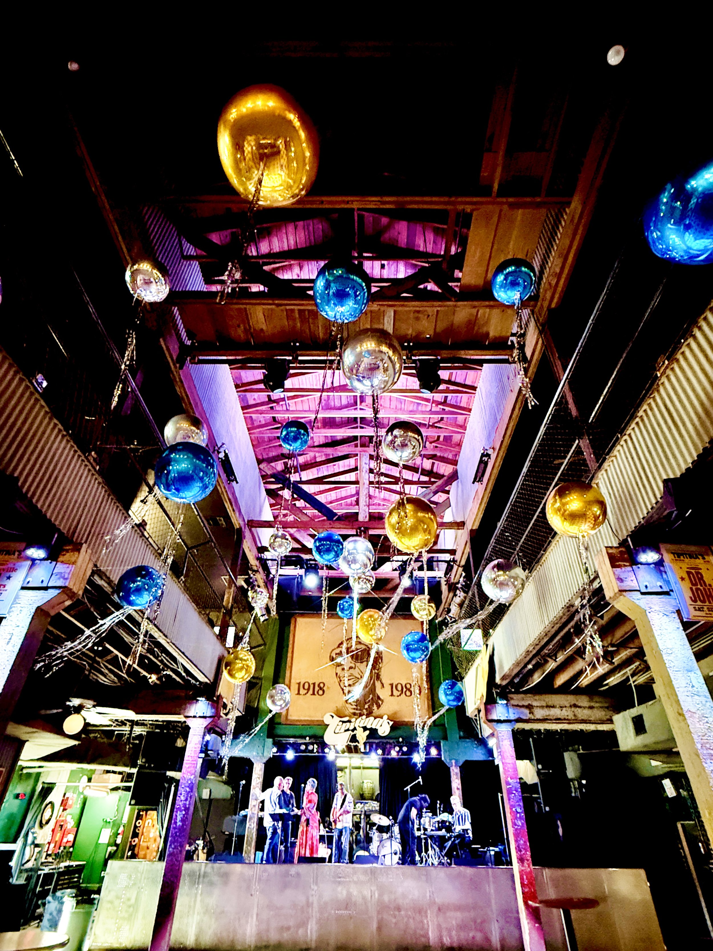 An indoor stage with a band performing, decorated with colorful hanging balloons, and a lit backdrop displaying the years 1918 and 198.