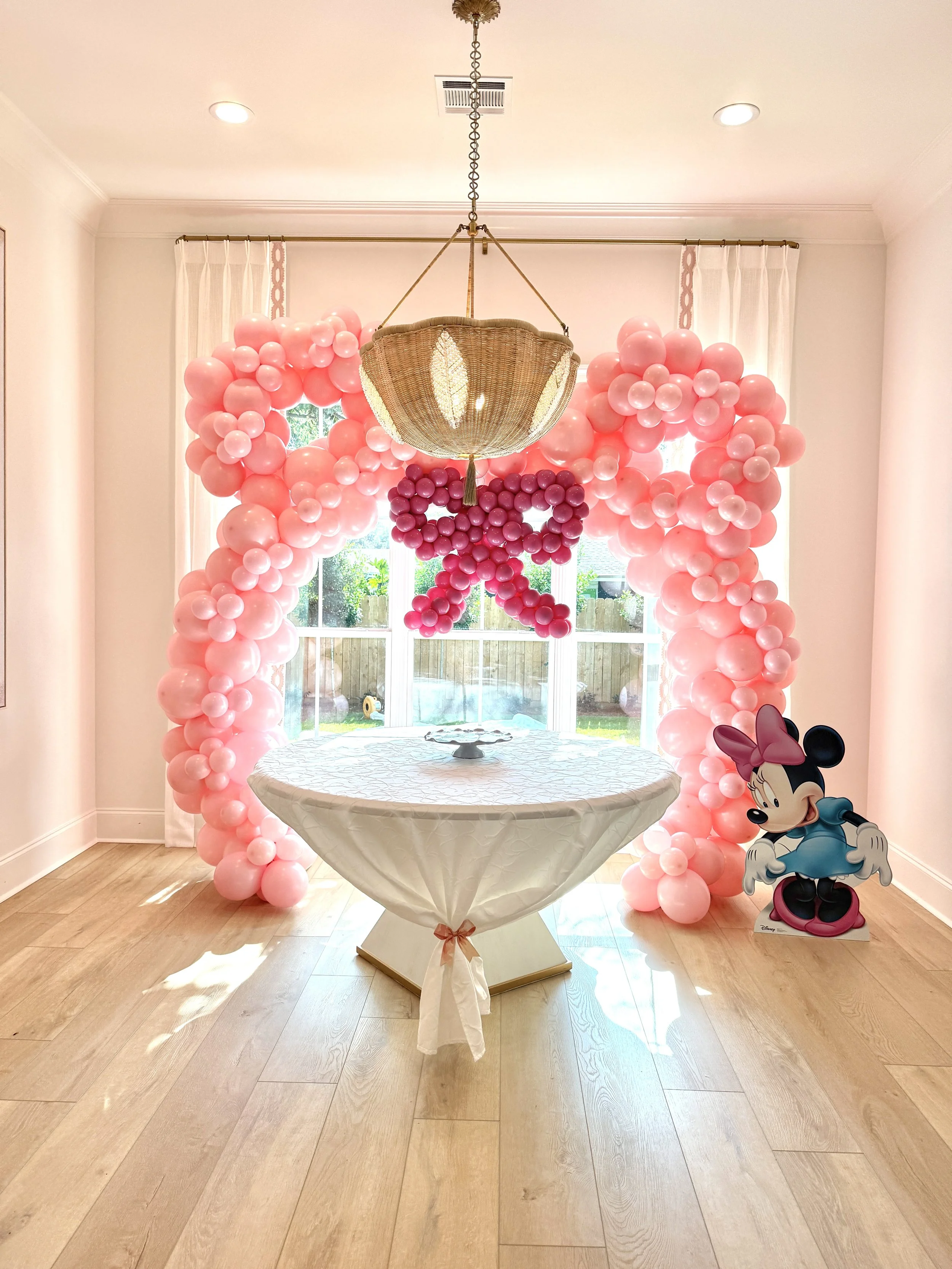 A room decorated for a celebration with pink balloon arch, a Minnie Mouse cardboard cutout, a table with a white tablecloth, and a window in the background.