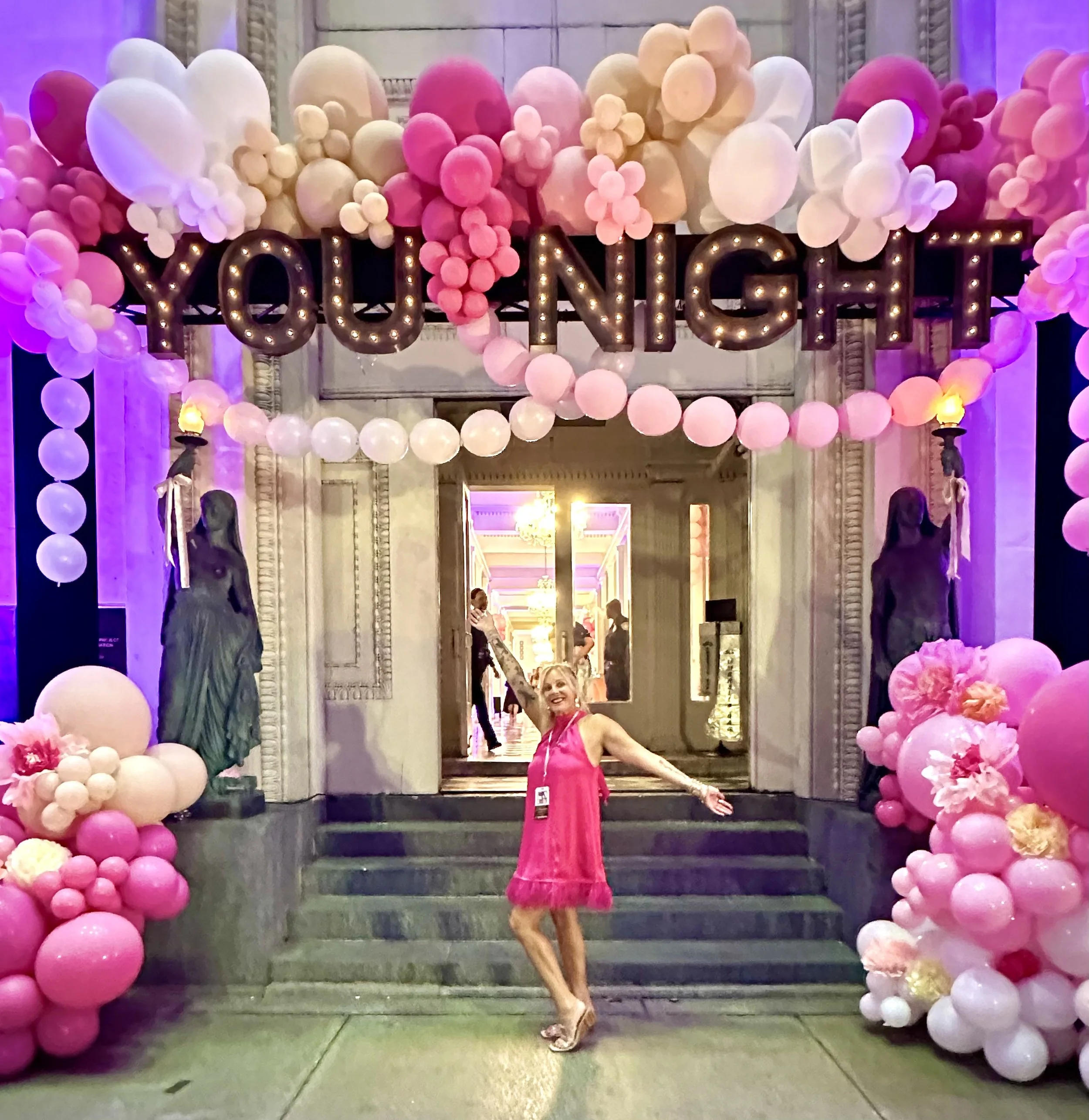 A woman in a pink dress standing on stairs decorated with pink, white, and cream balloons, with her arm raised and smiling.