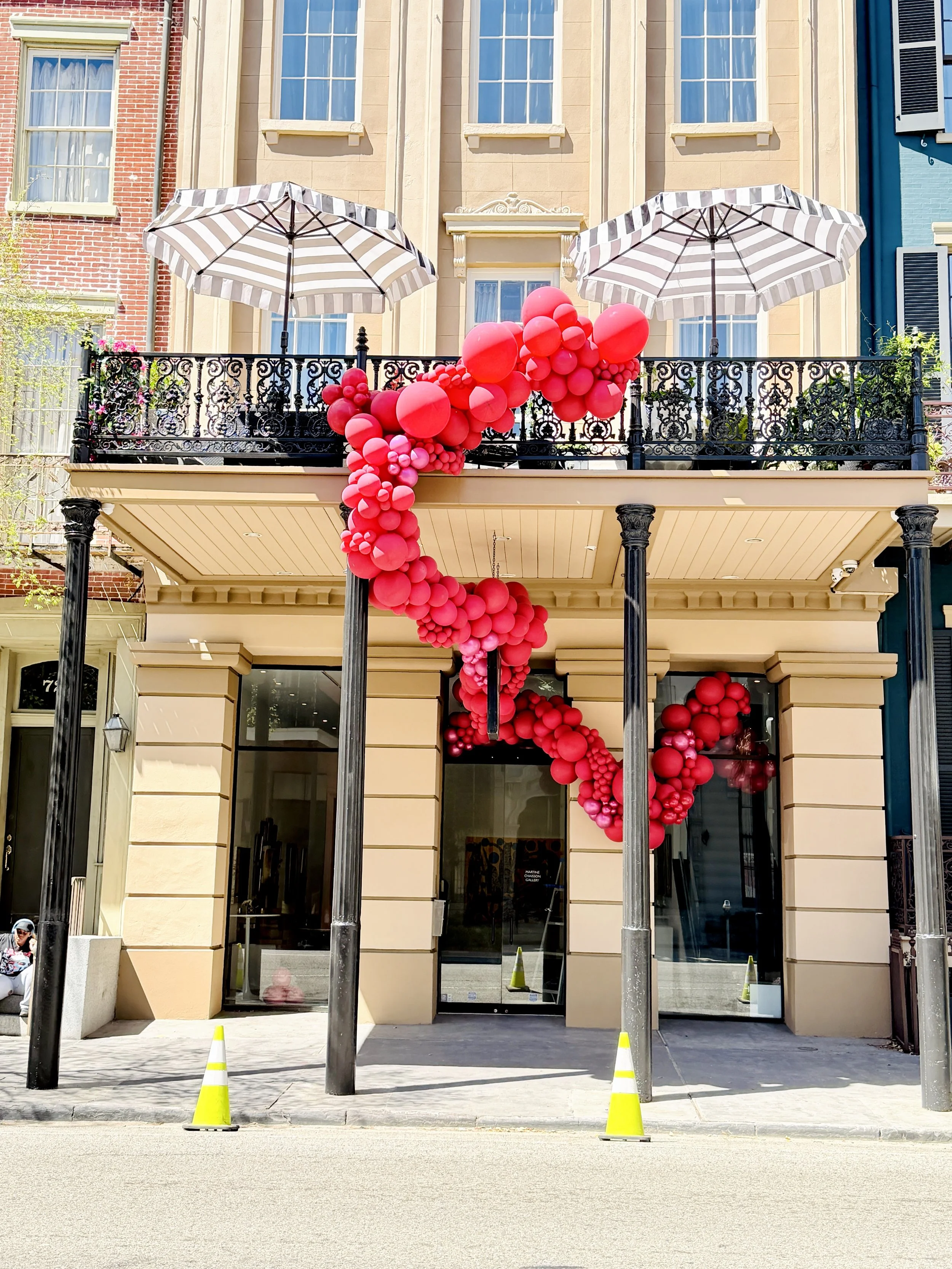 Storefront entrance decorated with red balloons arranged in a cascading pattern, with two black poles and yellow traffic cones on the sidewalk, and two striped umbrellas on the balcony above.