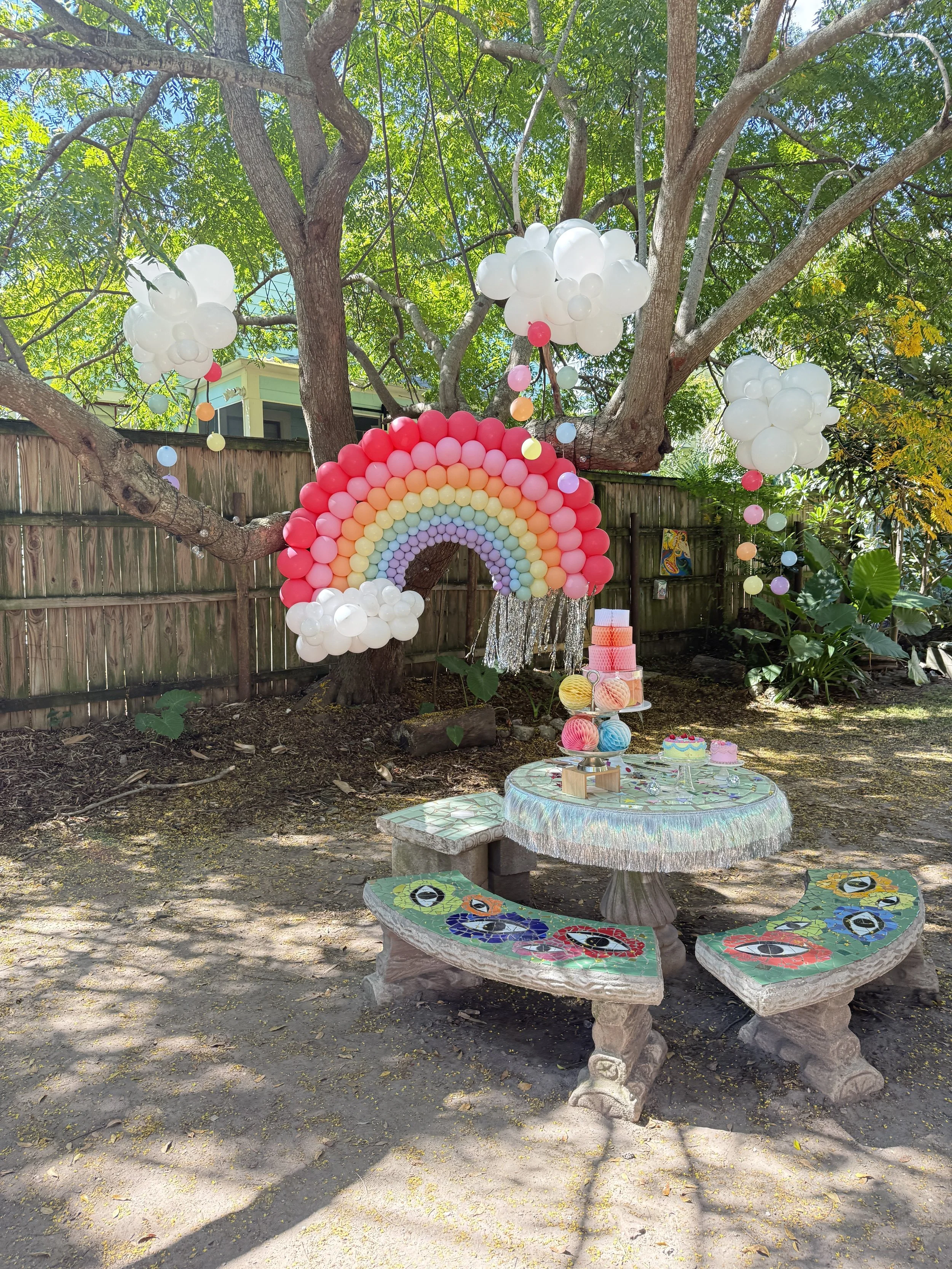 Decorated outdoor party setup featuring a balloon rainbow and clusters of balloons resembling clouds hanging from a tree. There are colorful table and bench settings on a stone table with mosaic patterns.