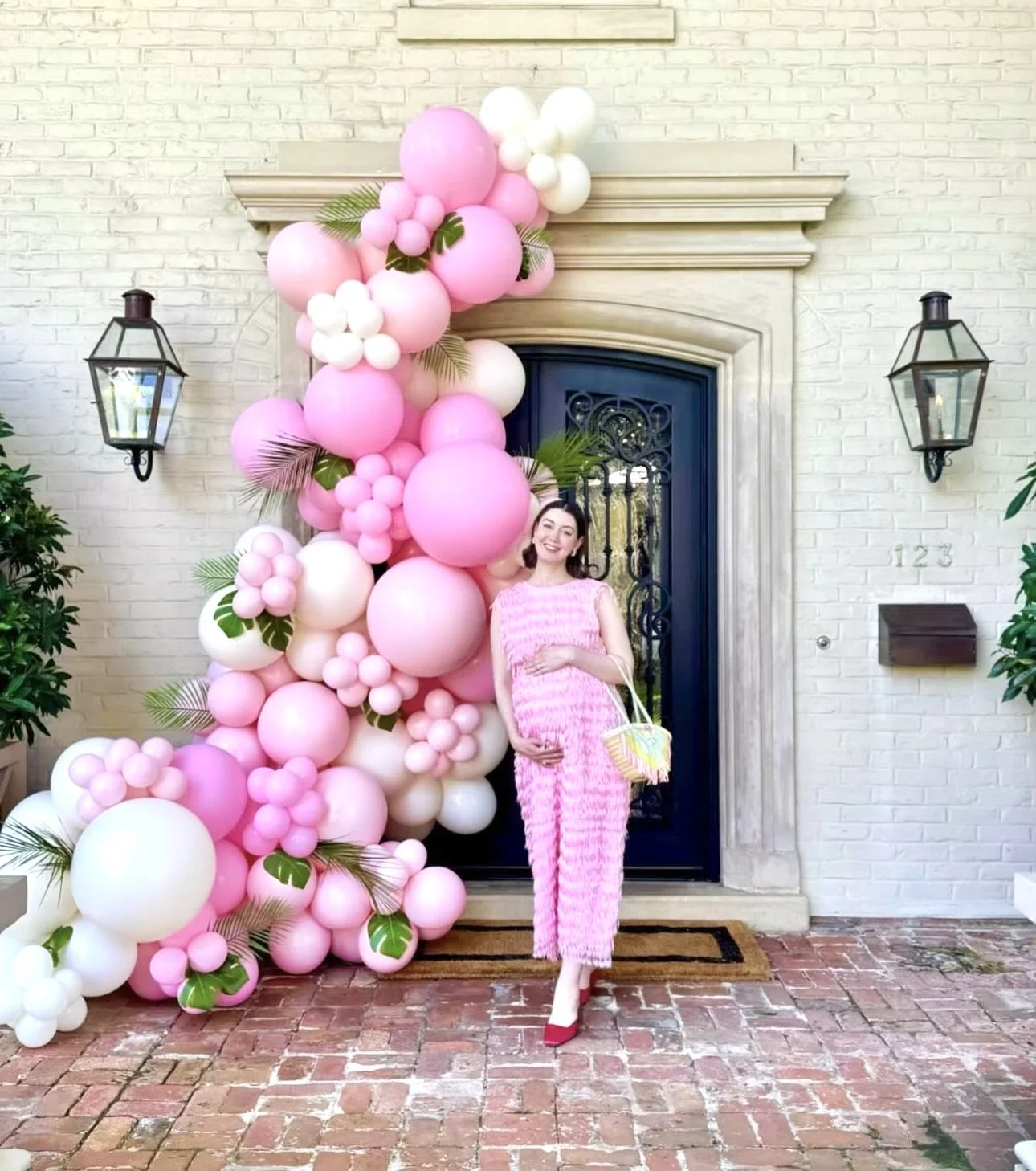 A pregnant woman in a pink dress stands in front of a decorative balloon arrangement in pink and white with green leaves outside a house with a brick pathway. She is holding her belly and a colorful basket.